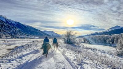 Zwei Reiter der Engadin River Ranch in Madulain (7523, Kanton Graubünden) reiten bei Sonnenschein durch eine verschneite Landschaft entlang des Inns mit Blick auf die umliegenden Berge.Zwei Reiter der Engadin River Ranch in Madulain (7523, Kanton Graubünden) reiten bei Sonnenschein durch eine verschneite Landschaft entlang des Inns mit Blick auf die umliegenden Berge.
