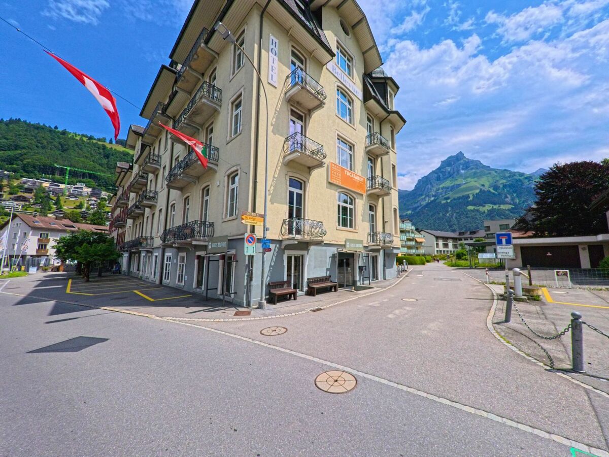 Ein mehrstöckiges Hotelgebäude in Engelberg (6390, Kanton Obwalden) steht an einer ruhigen Seitenstrasse, flankiert von wehenden Fahnen und mit Blick auf das markante Bergpanorama im Hintergrund.