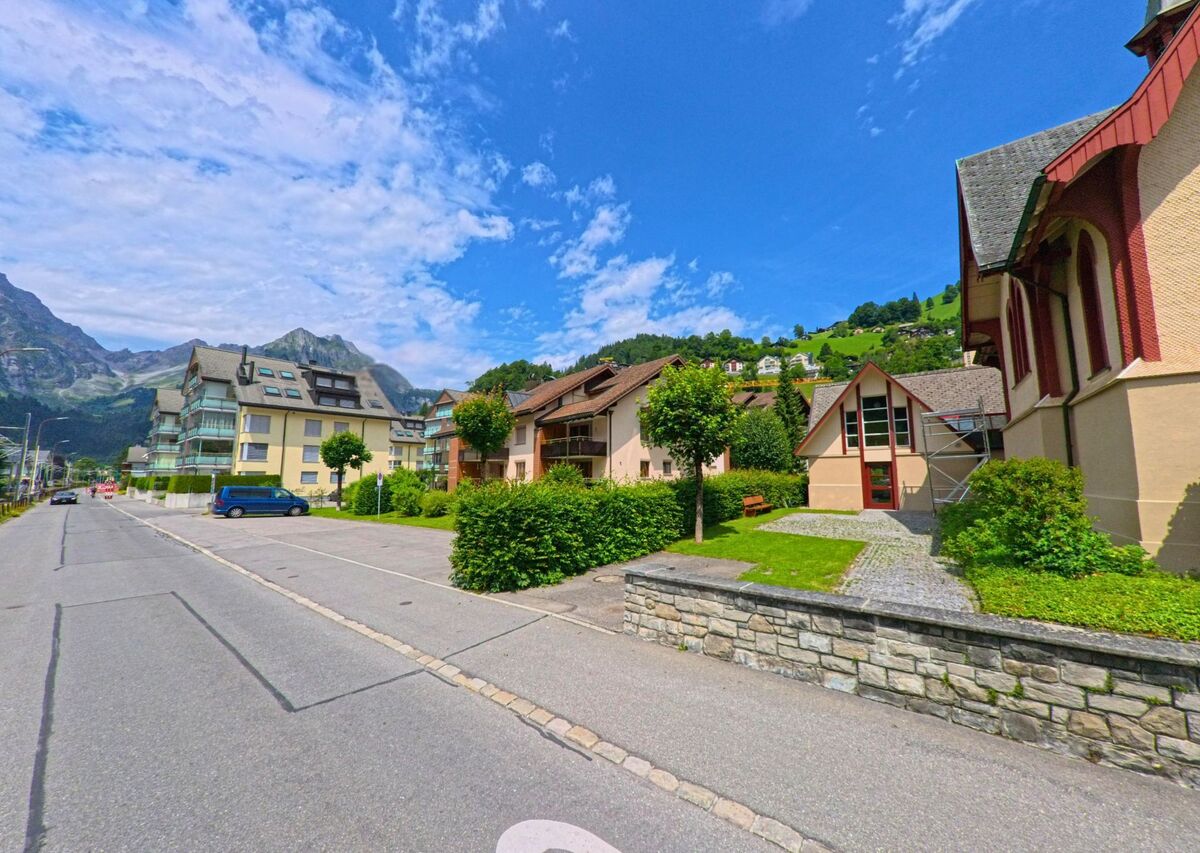 In Engelberg (6390, Kanton Obwalden) verlaufen Kirche und Wohnhaeuser entlang einer ruhigen Strasse mit Blick auf das Bergmassiv im Hintergrund.