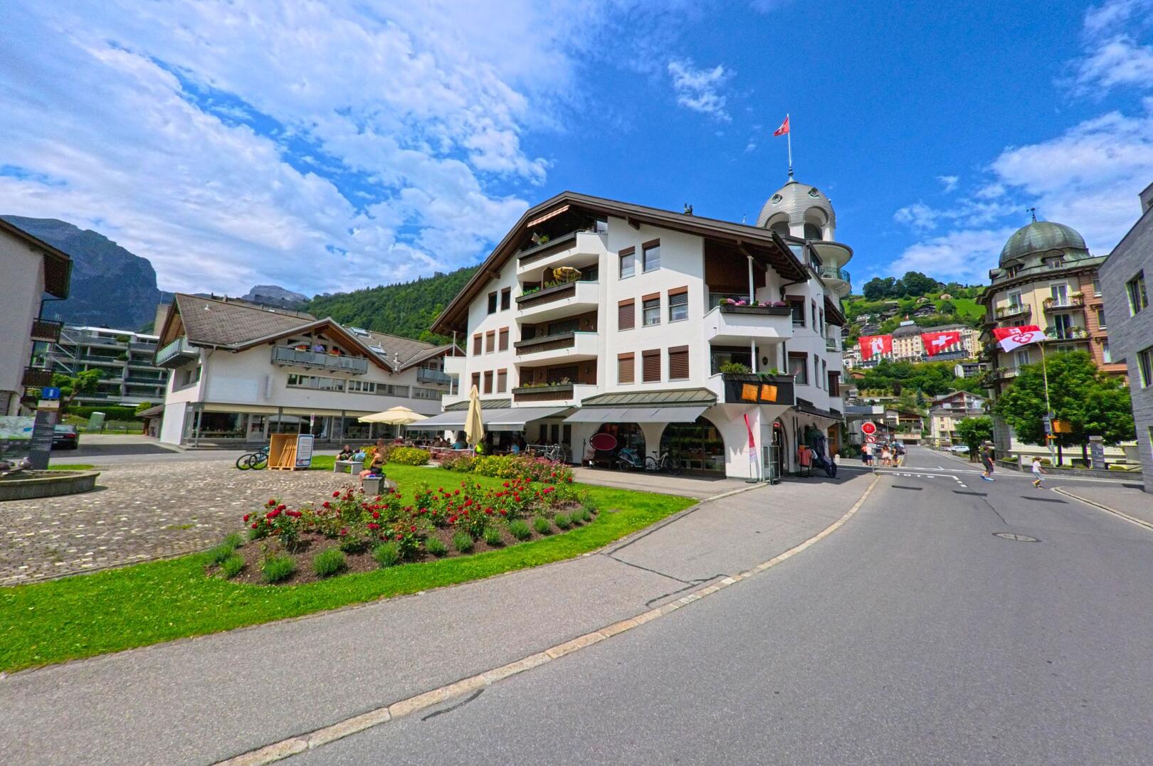 In Engelberg (6390, Kanton Obwalden) säumen moderne Wohnhäuser mit Schweizer Fahnen geschmückte Strassen, im Vordergrund blühen rote Rosen neben einem kleinen Platz mit Cafétischen.