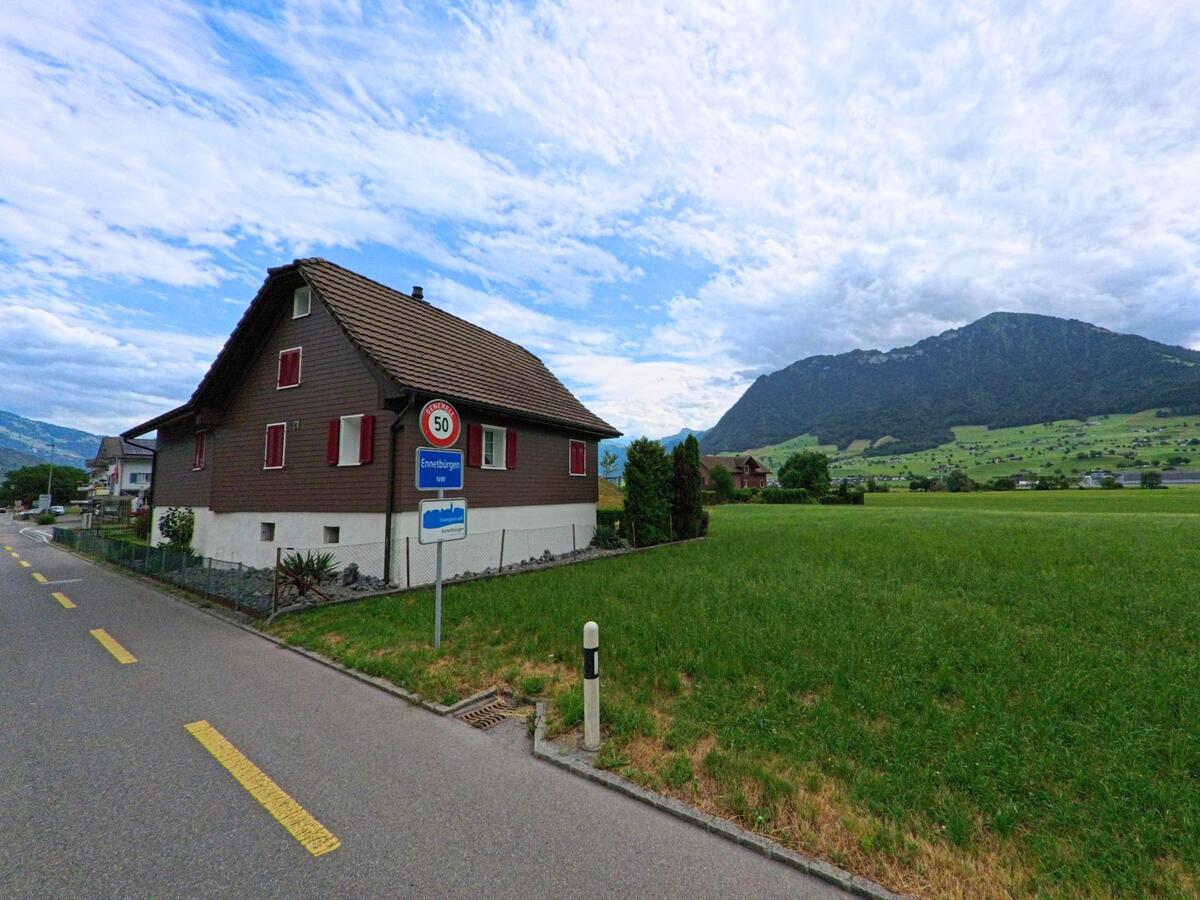 Am Ortseingang von Ennetbuergen (6373, Kanton Nidwalden) steht ein braunes Wohnhaus mit roten Fensterlaeden neben dem Ortsschild und weitem Blick auf gruene Felder und Huegel.