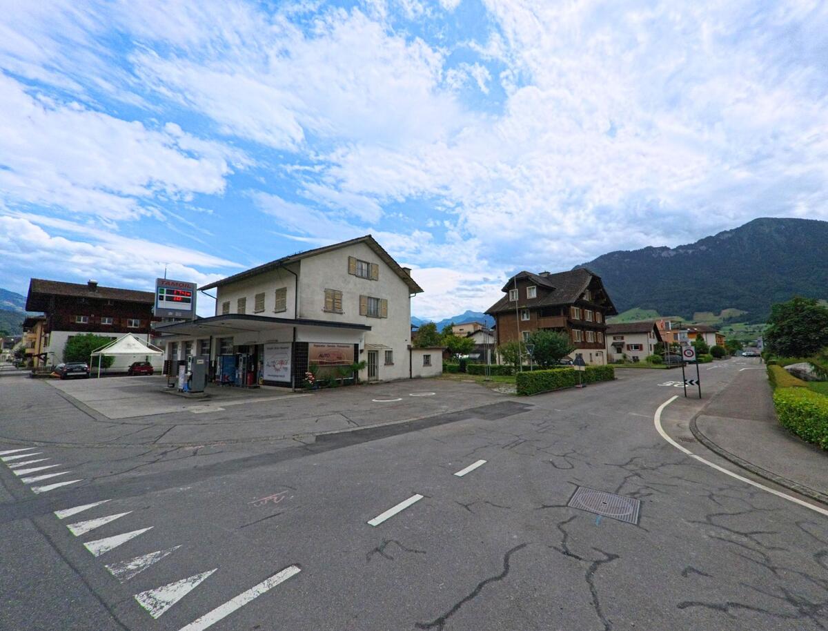 Eine kleine Tankstelle liegt in Ennetbuergen (6373, Kanton Nidwalden) an einer Kreuzung mit Wohnhaeusern und Blick auf bewaldete Huegel im Hintergrund.