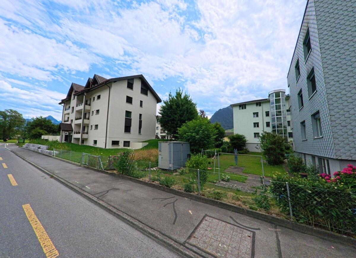 In Ennetbuergen (6373, Kanton Nidwalden) verlaufen Wohnhaeuser mit Gaerten und Wiesen entlang einer Nebenstrasse mit Blick auf bewaldete Huegel.