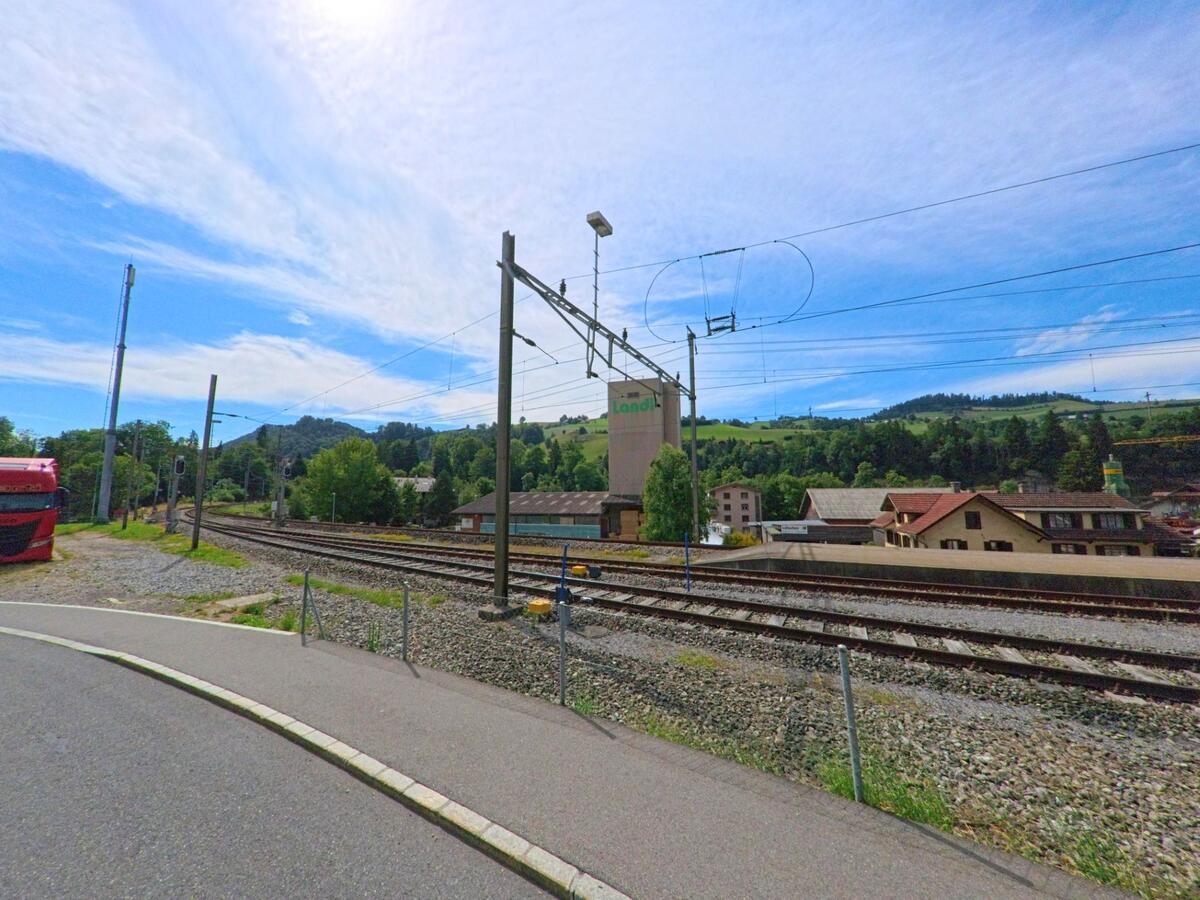 Die Bahngleise von Entlebuch (6162, Kanton Luzern) verlaufen unter freiem Himmel entlang von Wohnhäusern und einem markanten Lagergebäude mit Blick auf bewaldete Hügel.