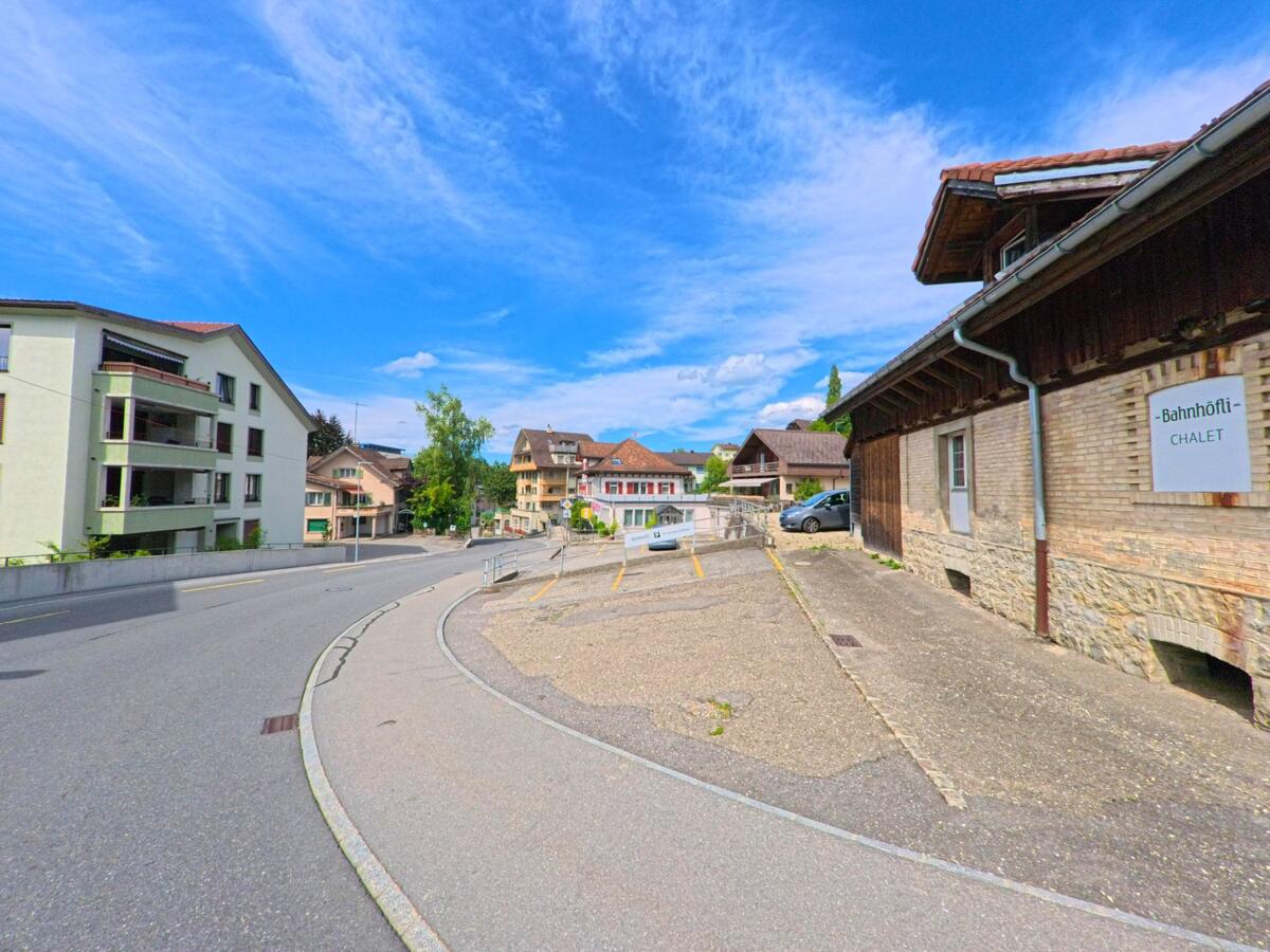  In Entlebuch (6162, Kanton Luzern) führt eine Strasse am ehemaligen Bahnhofsgebäude „Bahnhöfli-Chalet“ vorbei, flankiert von Wohnhäusern und Parkplätzen.