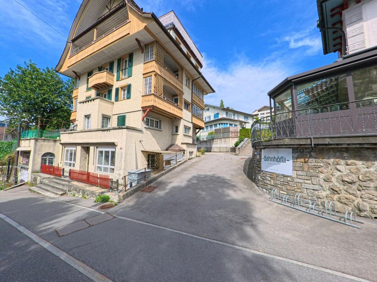  In Entlebuch (6162, Kanton Luzern) steht das Gebäude „Bahnhöfli“ mit markanter Veranda gegenüber einem traditionellen Wohnhaus mit Holzbalkonen an einer ansteigenden Strasse.