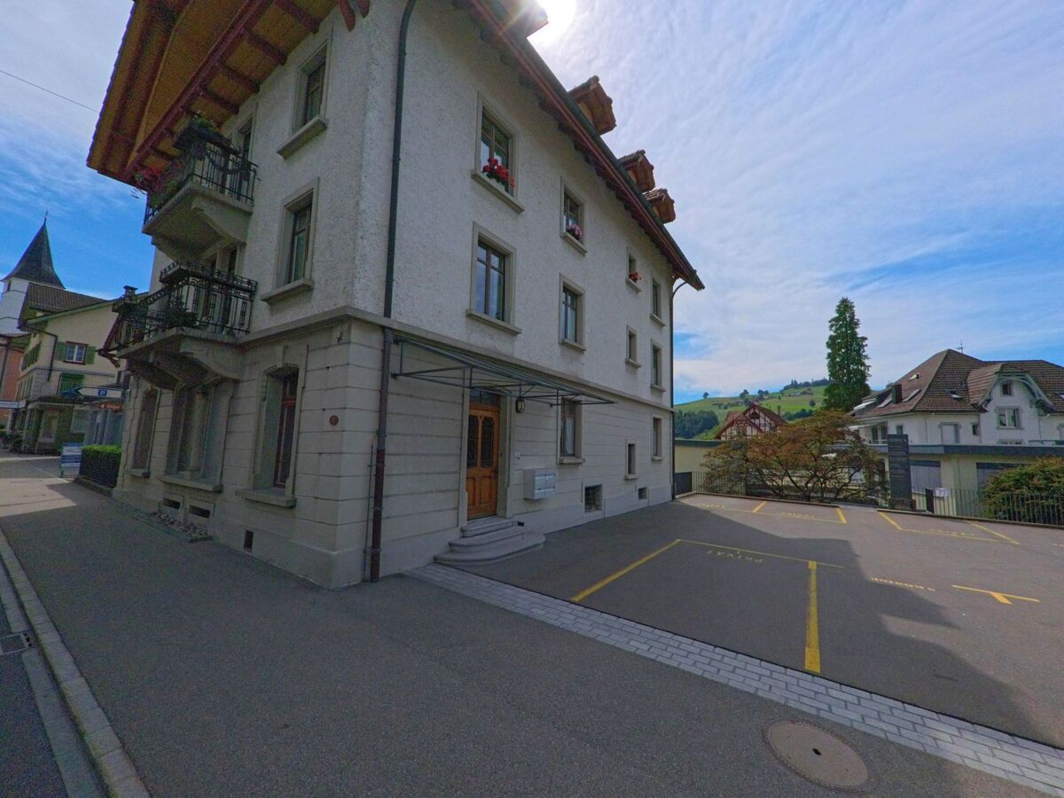  In Entlebuch (6162, Kanton Luzern) steht ein mehrstöckiges Wohnhaus mit Balkonen an einer ruhigen Seitenstrasse, dahinter zeigen sich grüne Hügel.