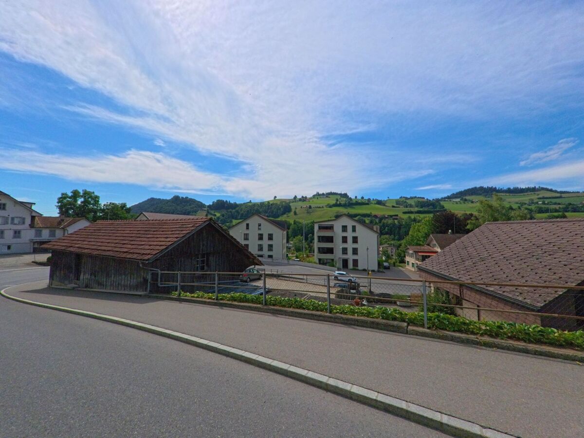 In Entlebuch (6162, Kanton Luzern) verläuft eine Nebenstrasse entlang traditioneller Holzschuppen mit Blick auf moderne Wohnhäuser und grüne Hügel im Hintergrund.