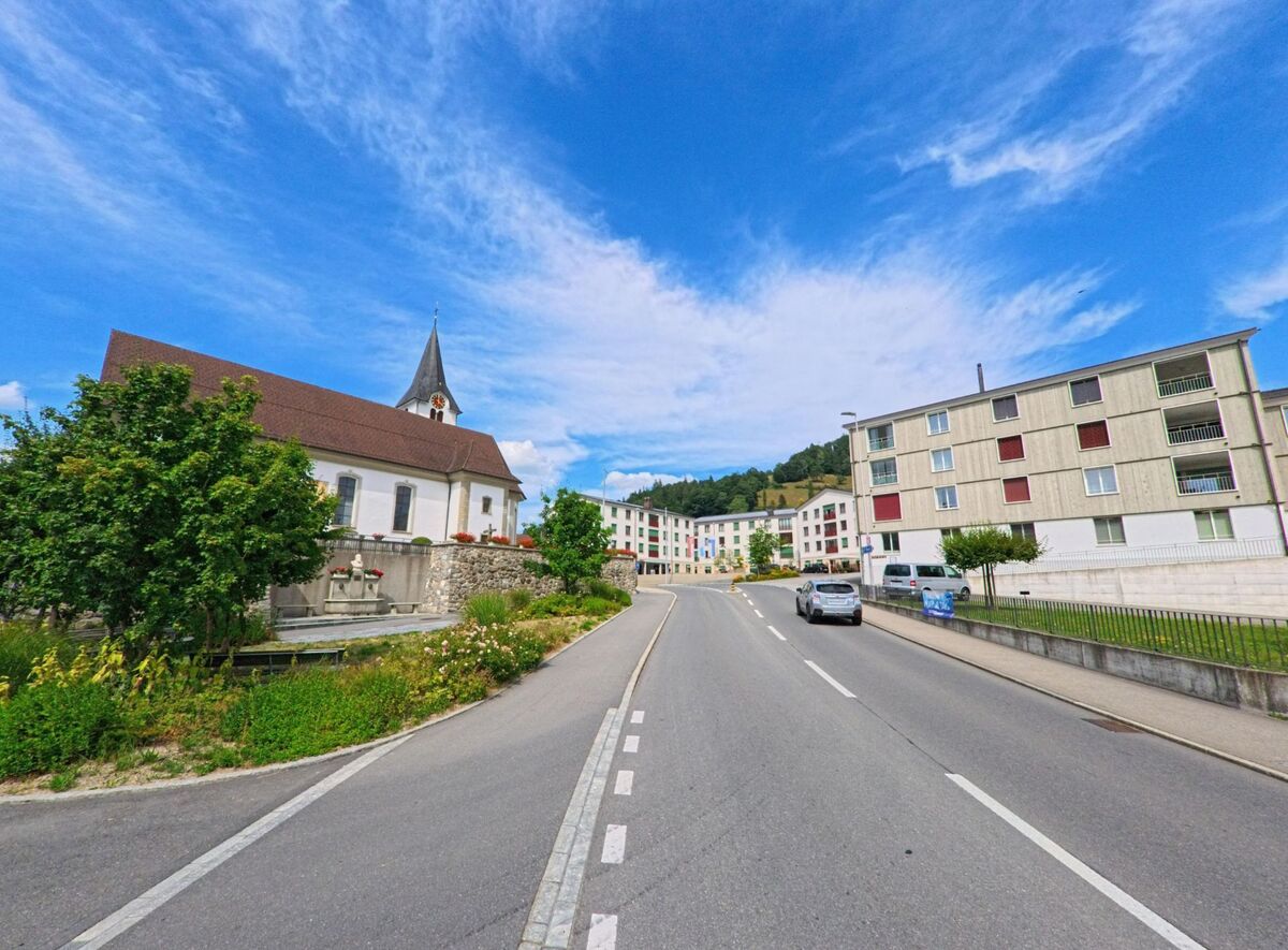 In Entlebuch (6162, Kanton Luzern) führt eine breite Strasse an der Kirche und modernen Wohnbauten vorbei in Richtung des Zentrums, umgeben von gepflegten Grünflächen.