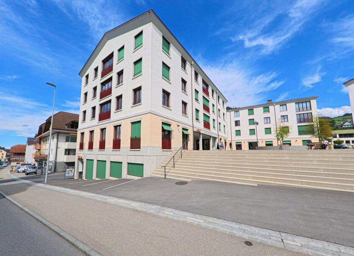 Eine breite Treppe führt in Entlebuch (6162, Kanton Luzern) zu modernen Wohn- und Geschäftsbauten mit farbigen Fensterläden und einem belebten Vorplatz.