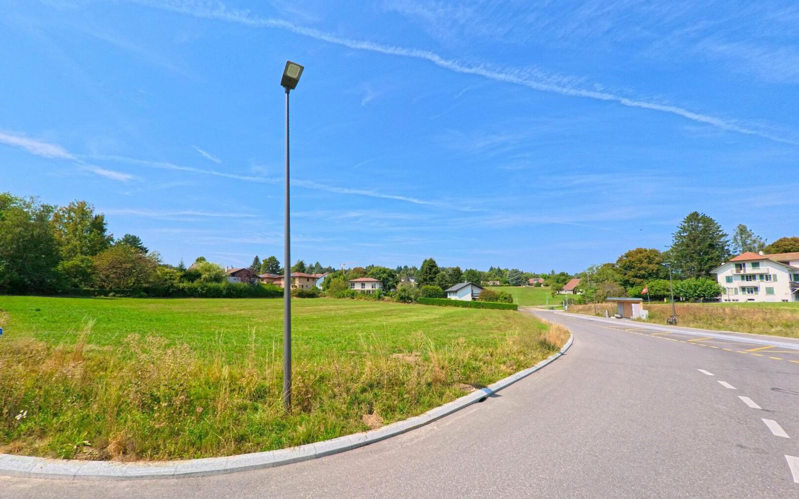 Am Ortsrand von Épalinges (1066, Kanton Waadt) liegt eine breite Gruenflaeche neben der Strasse, im Hintergrund sind einzelne Wohnhaeuser sichtbar.
