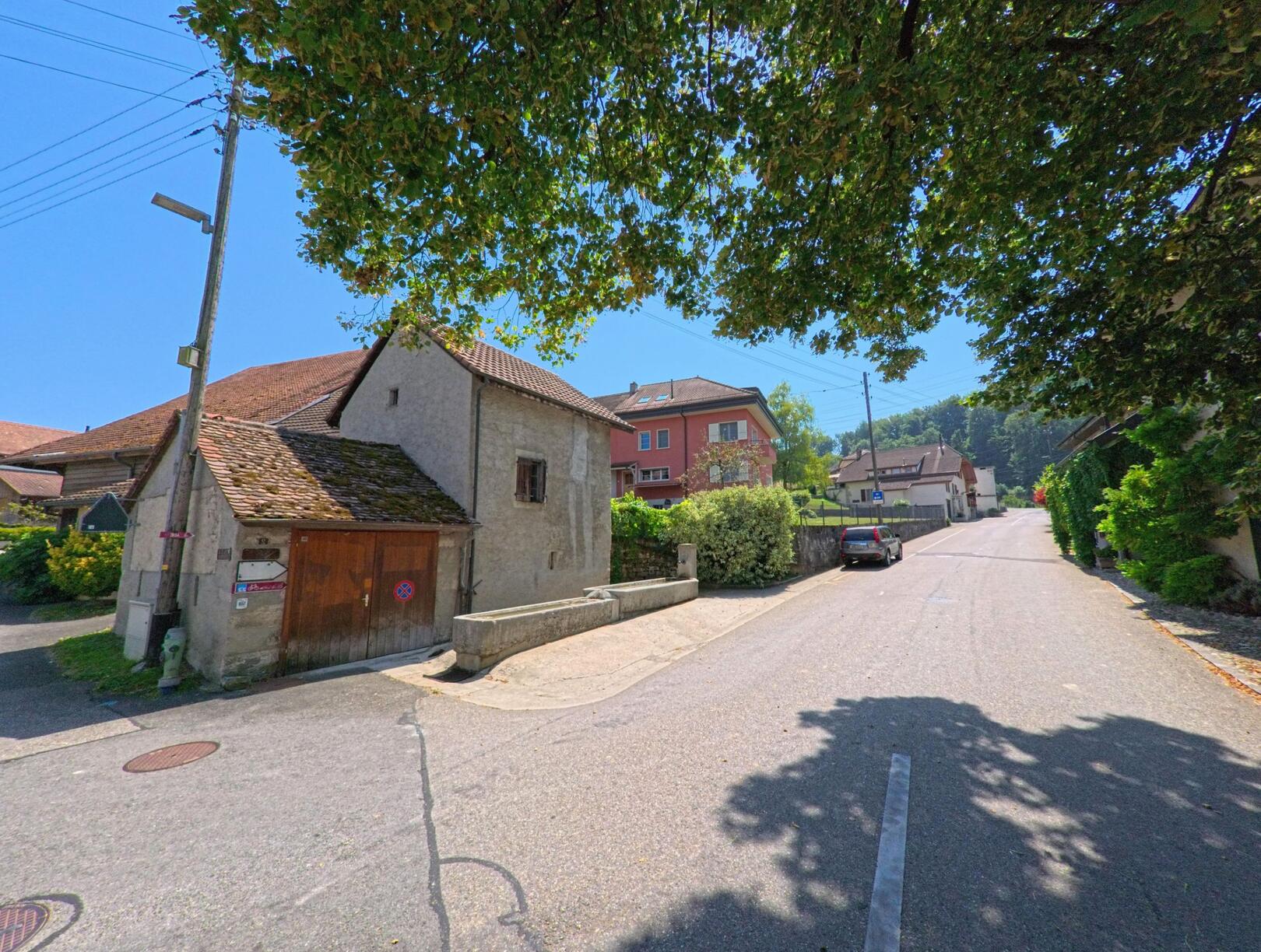 In Ependes (1434, Kanton Waadt) fuehrt eine leicht ansteigende Strasse vorbei an traditionellen Wohnhaeusern und einem alten Gebaeude mit Holztor.