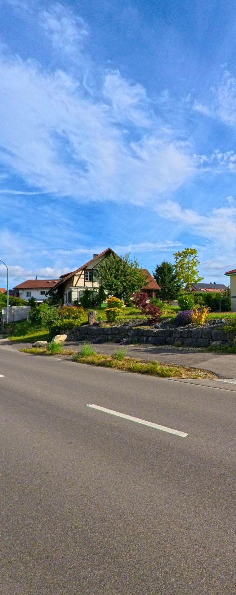 Vor einem bunt bepflanzten Garten erhebt sich in Epsach (3272, Kanton Bern) ein traditionelles Wohnhaus, das direkt an einer vorbeifuehrenden Strasse liegt.