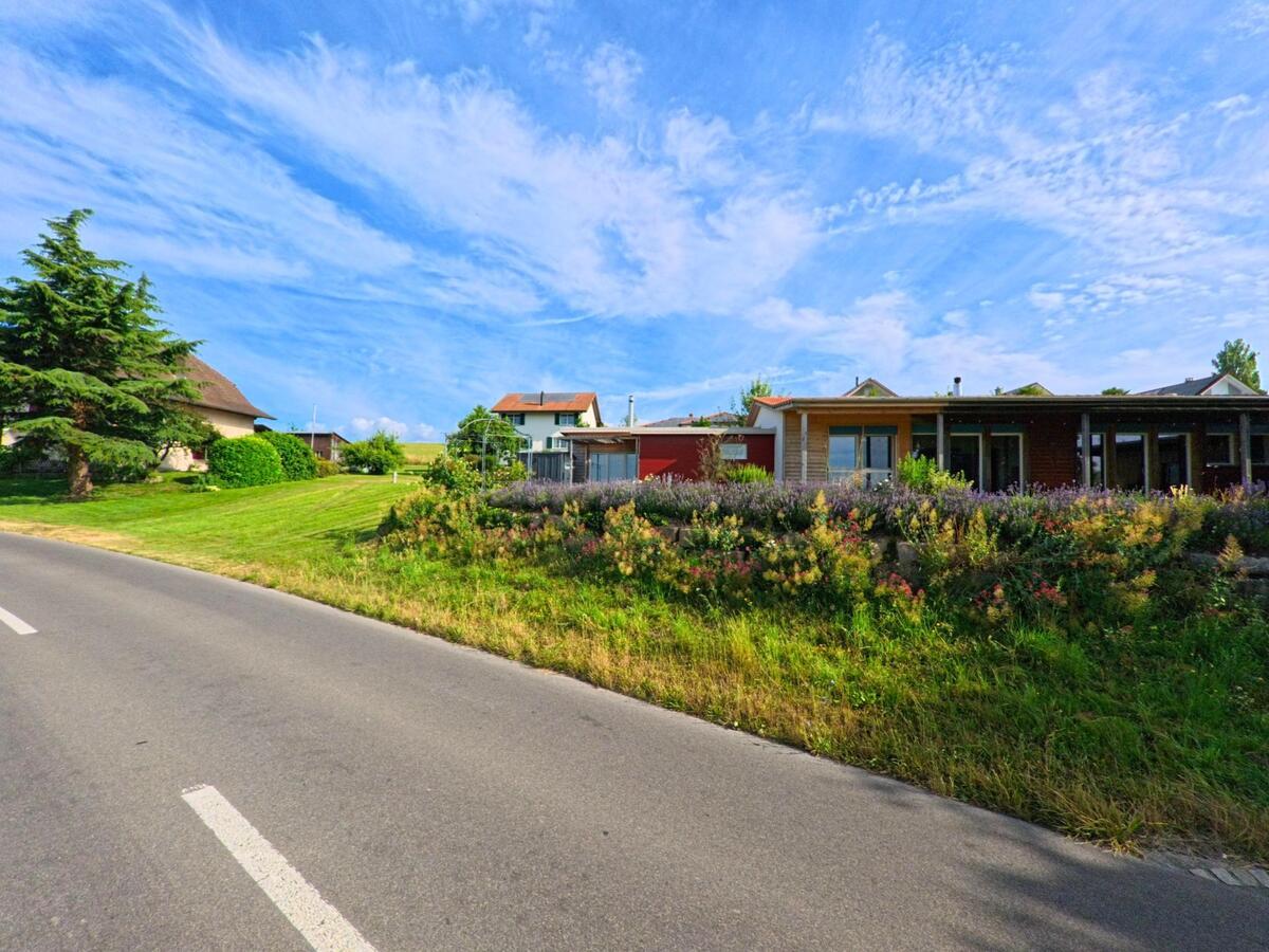 Entlang der Strasse in Epsach (3272, Kanton Bern) liegen gepflegte Gärten und Wohnhäuser, die von bunten Pflanzen und einem breiten Grünstreifen begleitet werden.