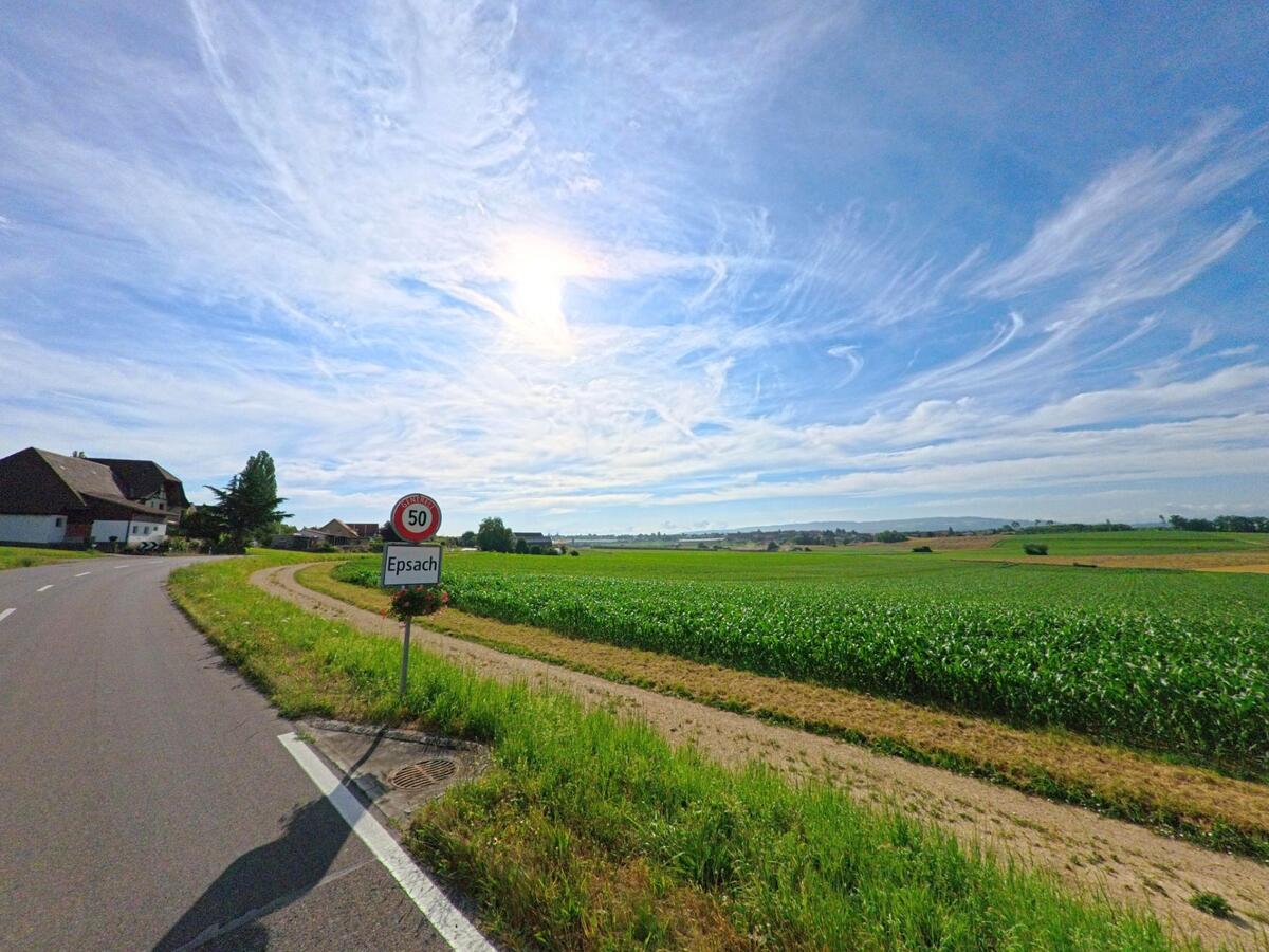 Am Ortseingang von Epsach (3272, Kanton Bern) steht ein Geschwindigkeitsschild vor weiten Feldern, waehrend die Strasse sanft ins Dorf fuehrt.