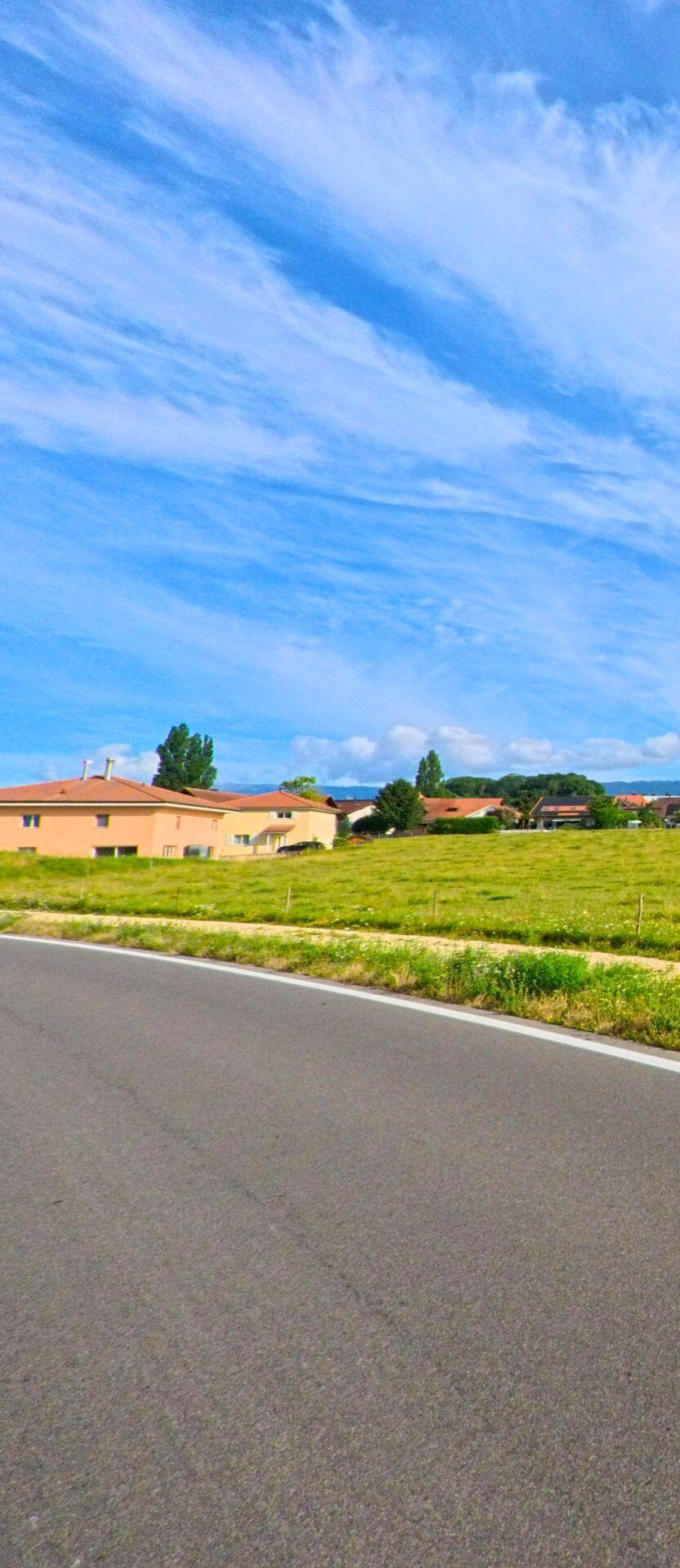 Hinter einer sanft geschwungenen Strasse in Epsach (3272, Kanton Bern) liegen Wiesen und mehrere Wohnhaeuser, die sich vor dem weiten Himmel deutlich abzeichnen.