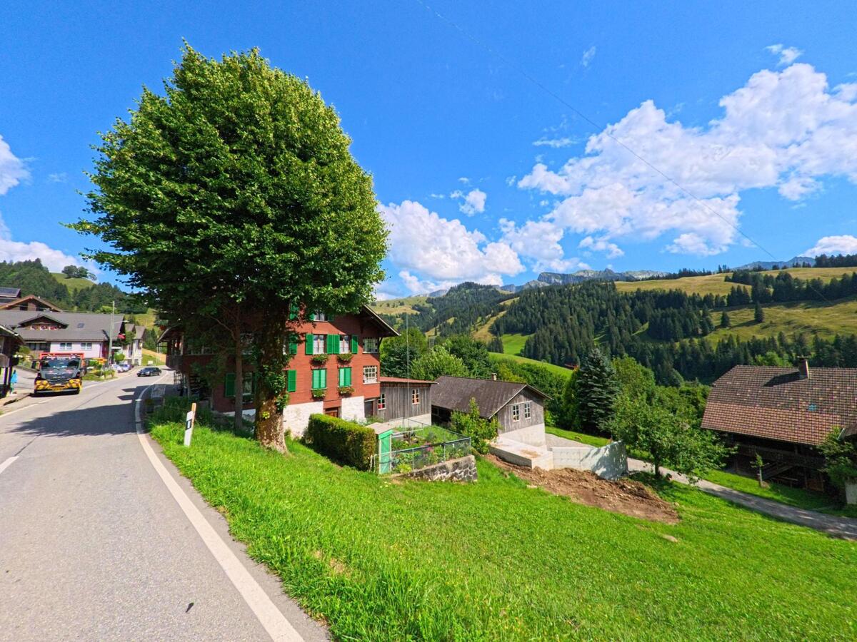 Am Ortseingang von Eriz (3619, Kanton Bern) steht ein rotes Wohnhaus mit grünen Fensterlaeden neben einer Landstrasse, waehrend sich im Hintergrund bewaldete Huegel und Almweiden erheben.
