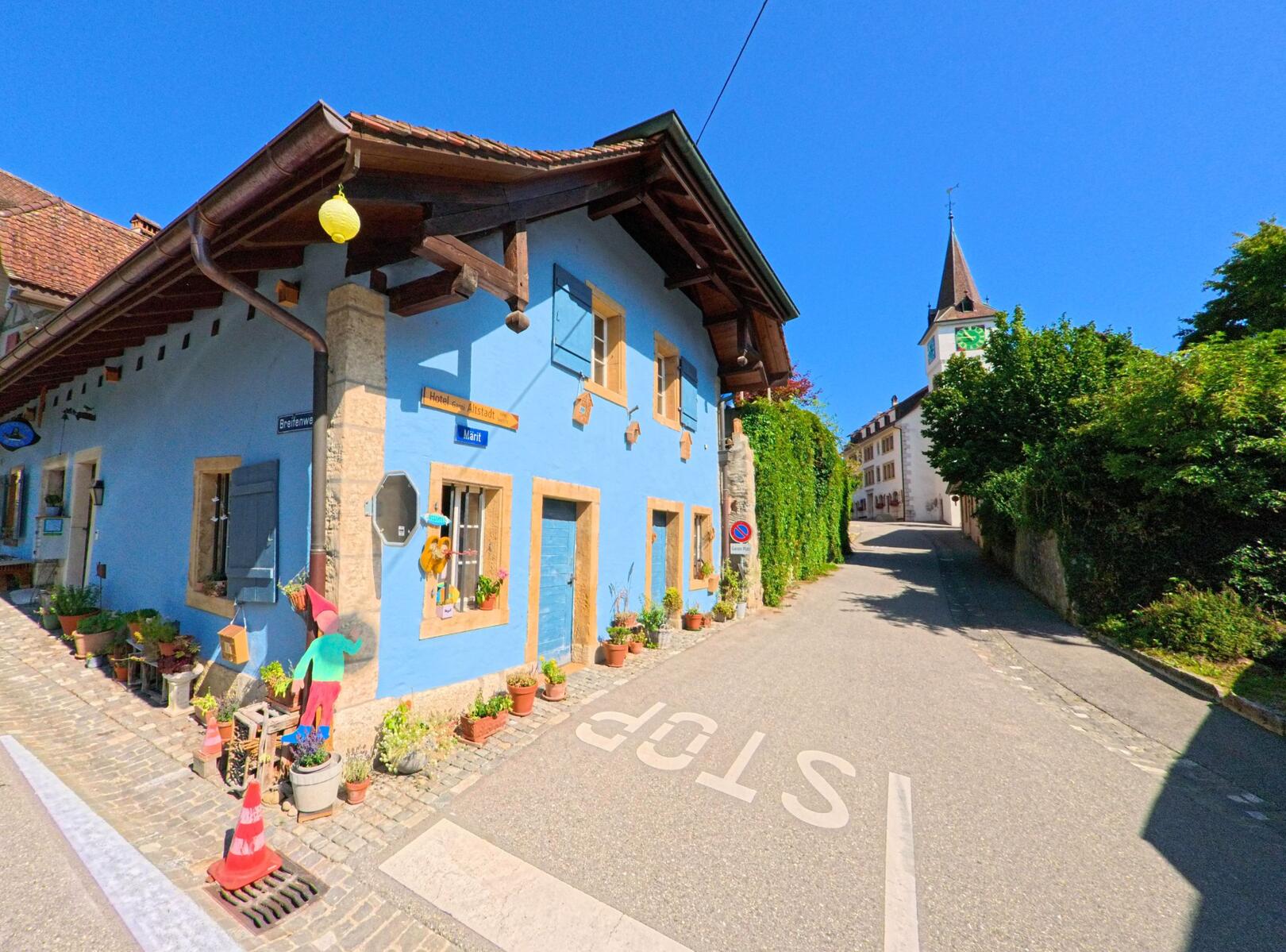 In der Altstadt von Erlach (3235, Kanton Bern) steht ein blau gestrichenes Haus mit Blumenschmuck an den Fenstern, waehrend im Hintergrund der Kirchturm ueber einer ansteigenden Gasse sichtbar ist.
