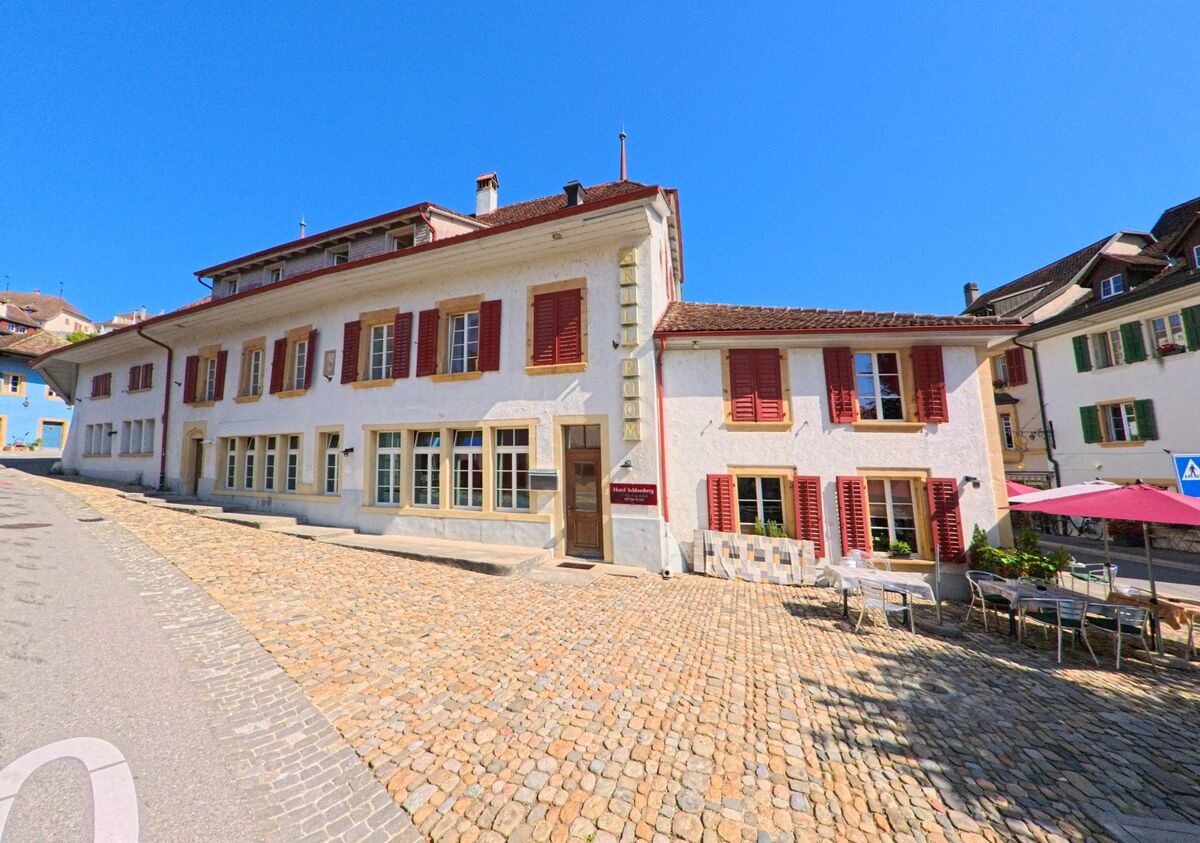 An einem gepflasterten Platz in Erlach (3235, Kanton Bern) steht ein helles Gebaeude mit roten Fensterlaeden, vor dem Tische und Stuehle eines Strassencafes aufgestellt sind.