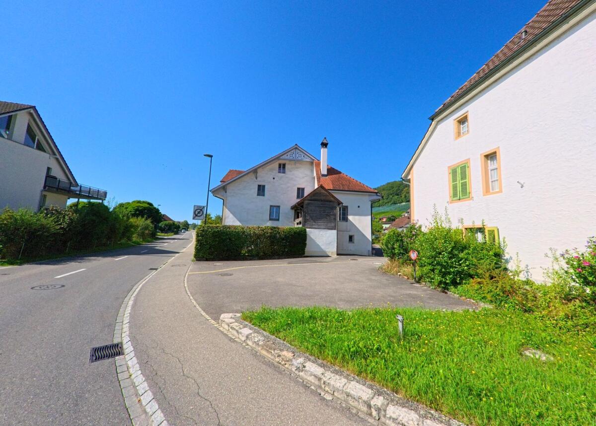 Eine kurvige Strasse fuehrt durch ein Wohngebiet in Erlach (3235, Kanton Bern), gesaeumt von hellen Haeusern mit farbigen Fensterlaeden und gepflegten Gaerten.