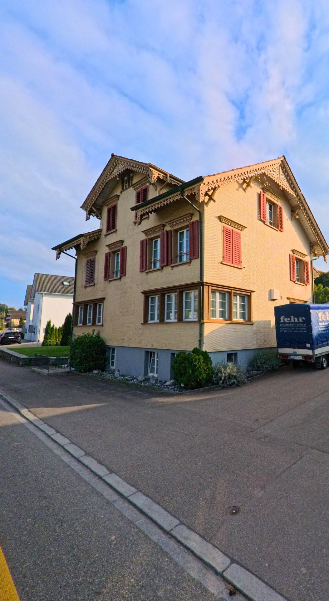 Ein mehrstöckiges Wohnhaus mit geschnitzten Dachdetails und roten Fensterläden steht in Erlen (8586, Kanton Thurgau) an einer ruhigen Seitenstrasse.