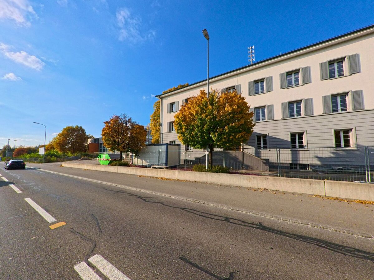 Ein grosses Gebäude mit hellen Fensterläden steht in Erlen (8586, Kanton Thurgau) an einer Strasse, begleitet von herbstlich gefärbten Bäumen und einem Trottoir.