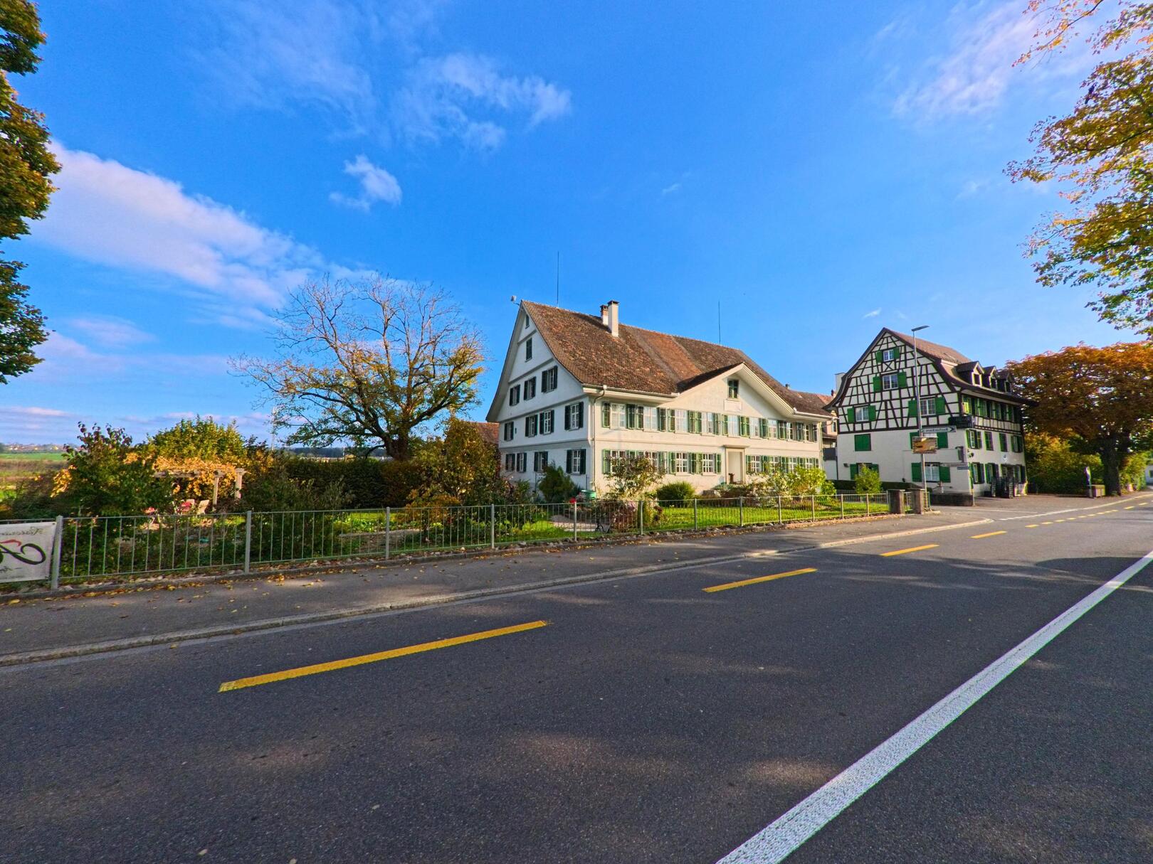 An einer Strasse in Erlen (8586, Kanton Thurgau) stehen zwei traditionelle Wohnhäuser mit gepflegtem Garten unter blauem Himmel.