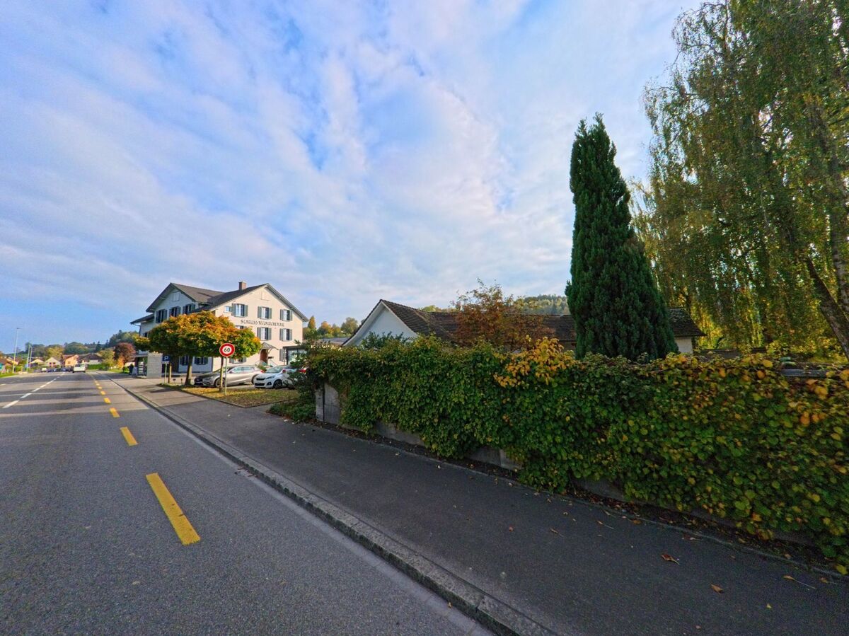 An einer Hauptstrasse in Erlen (8586, Kanton Thurgau) führt ein Trottoir entlang einer begrünten Mauer mit Bäumen, während im Hintergrund ein grosses Gebäude und Parkplätze zu sehen sind.