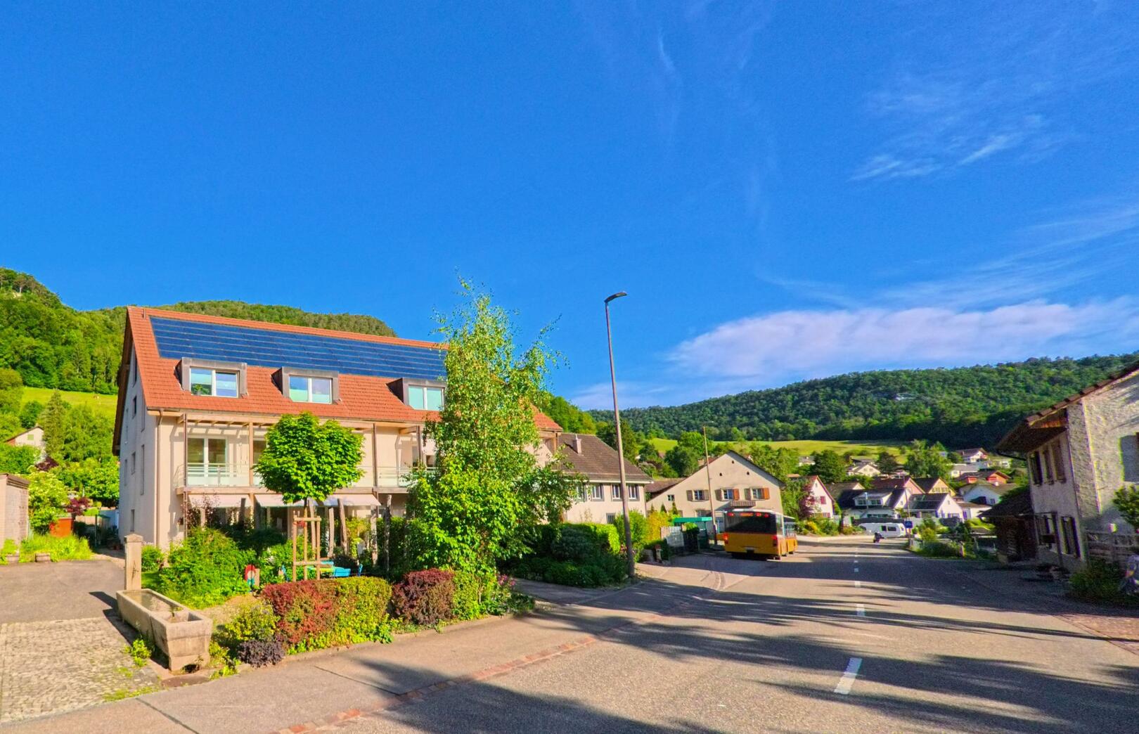 In Erschwil (4228, Kanton Solothurn) säumen Einfamilienhäuser mit Solaranlagen eine ruhige Dorfstrasse, während ein gelber Bus Richtung Zentrum fährt und grüne Hügel den Hintergrund bilden.