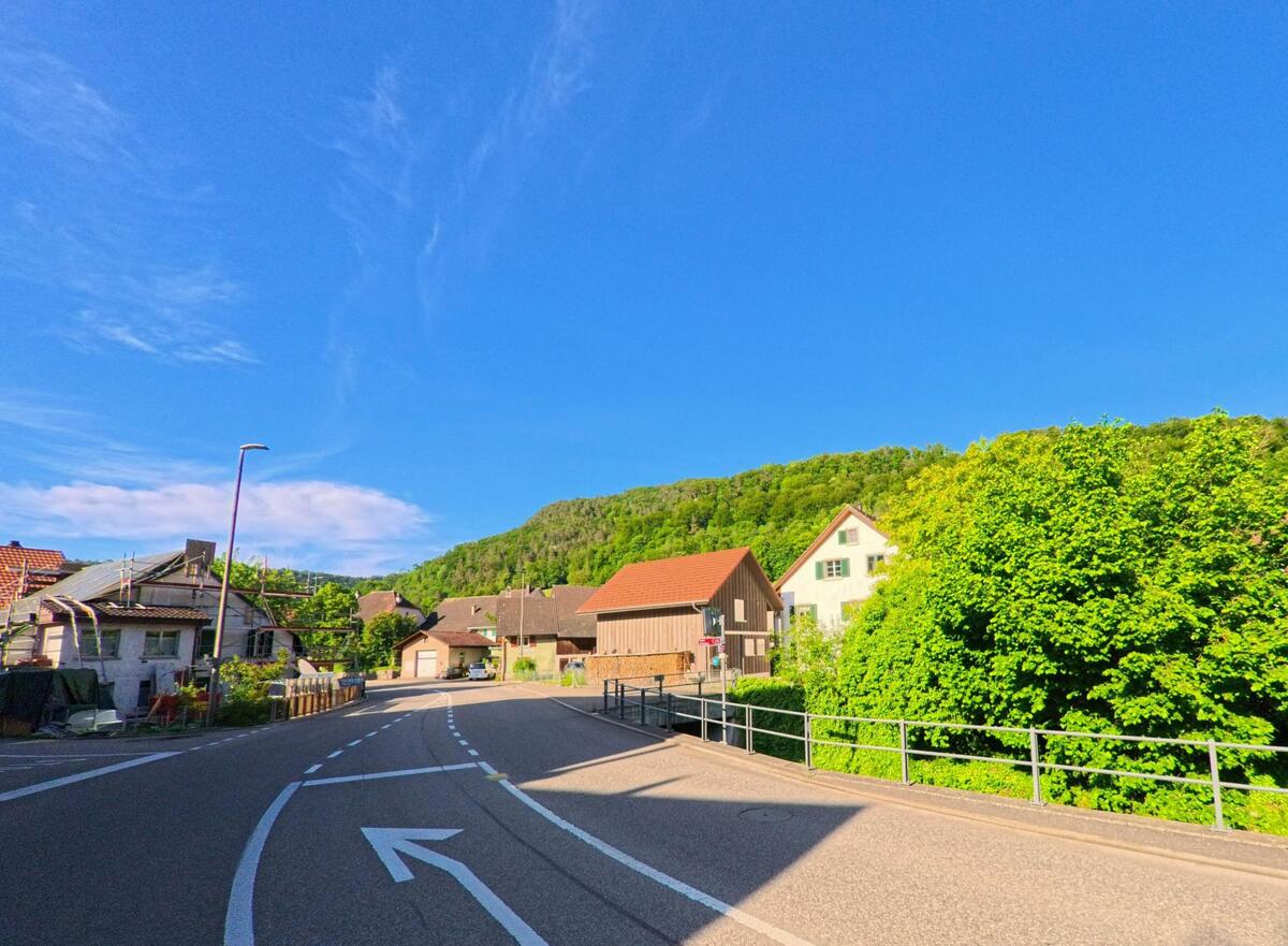 In Erschwil (4228, Kanton Solothurn) windet sich eine Strasse mit klarer Fahrbahnmarkierung durch ein Wohngebiet mit traditionellen Häusern und grün bewachsenen Hängen im Hintergrund.