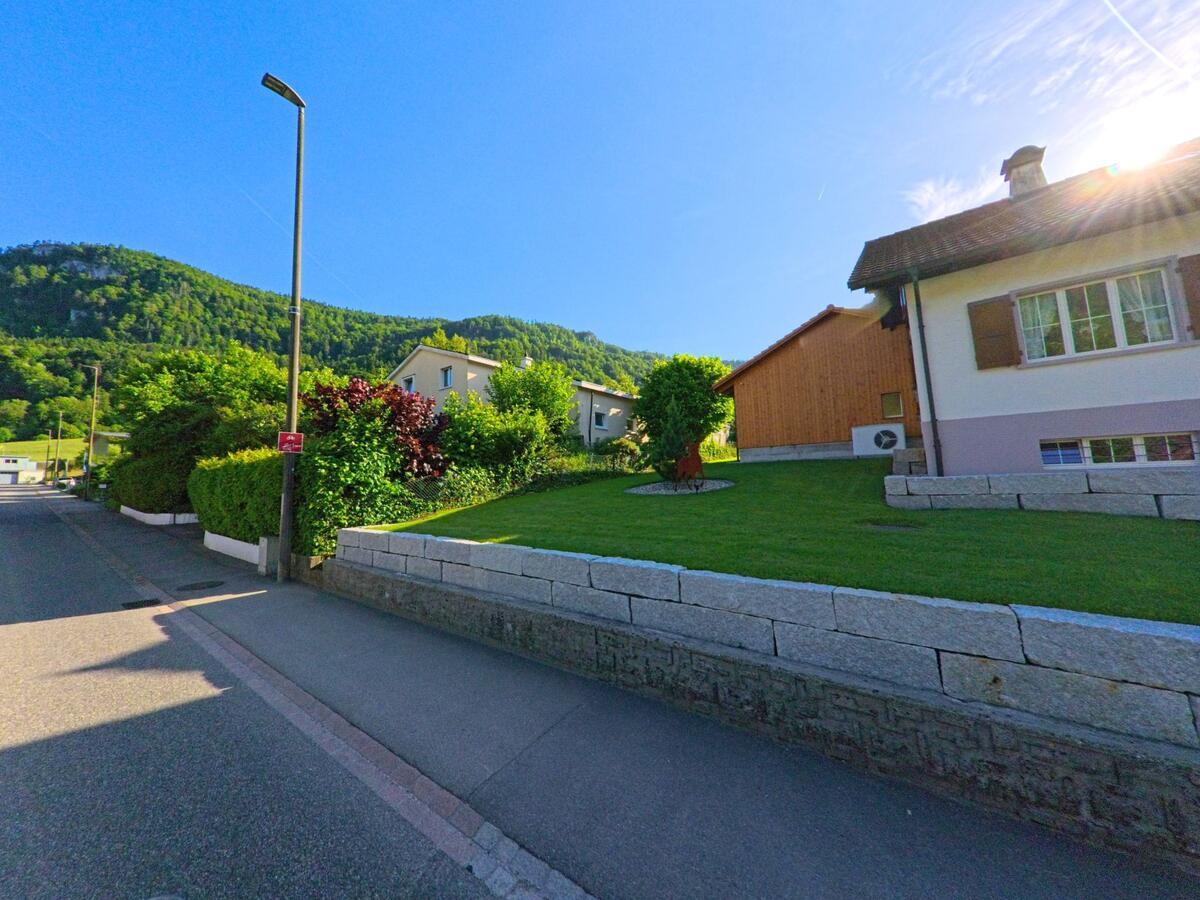  In Erschwil (4228, Kanton Solothurn) führt eine ruhige Quartierstrasse an gepflegten Einfamilienhäusern mit Vorgärten vorbei, eingerahmt von bewaldeten Hügeln im Hintergrund.
