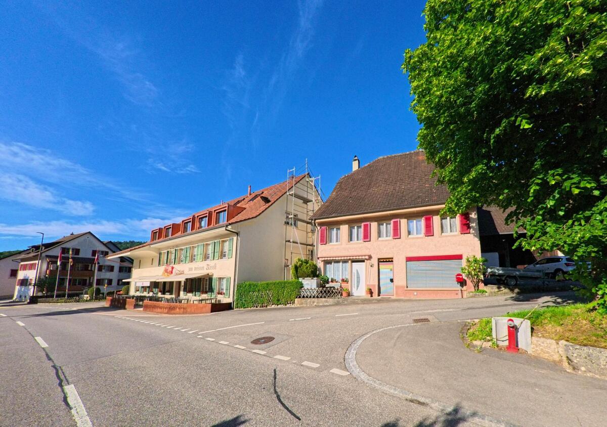 Im Zentrum von Erschwil (4228, Kanton Solothurn) befindet sich das Restaurant „Zum Weissen Rössli“ an einer belebten Strassenecke, flankiert von Wohnhäusern mit farbigen Fensterläden.
