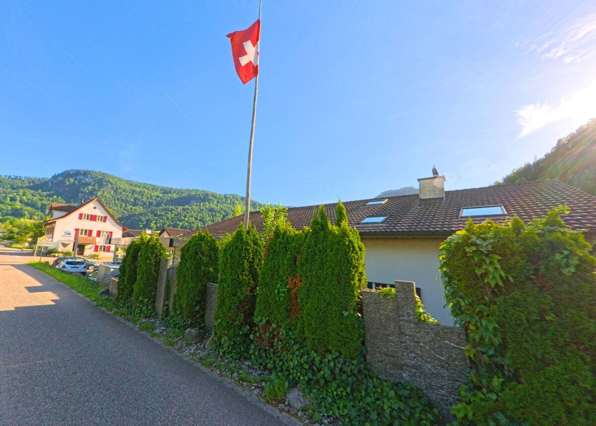 In Erschwil (4228, Kanton Solothurn) weht eine Schweizer Fahne über einem Wohnquartier mit gepflegten Hecken und Blick auf bewaldete Hügel im Hintergrund.
