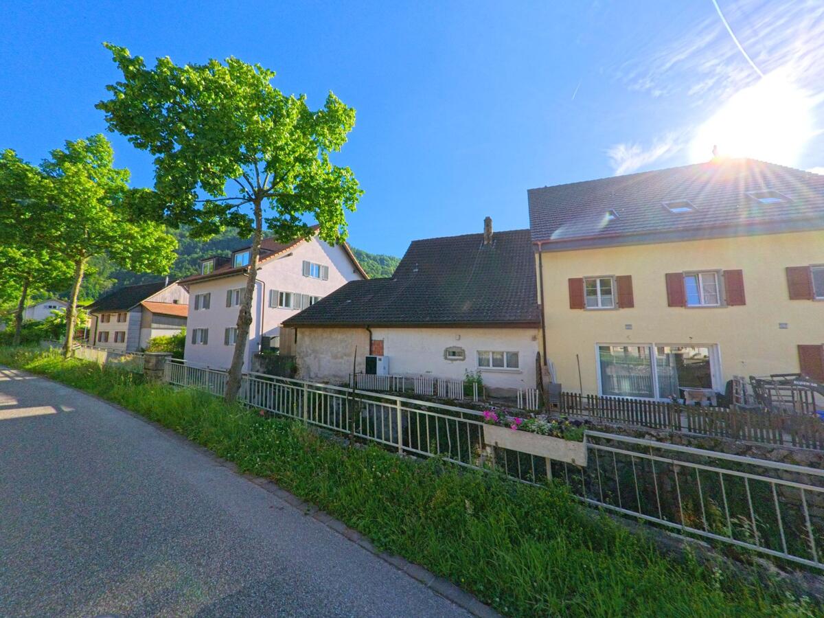  In Erschwil (4228, Kanton Solothurn) verläuft eine schmale Strasse an einem offenen Wasserlauf mit Geländer entlang, gesäumt von Wohnhäusern und schattenspendenden Bäumen.