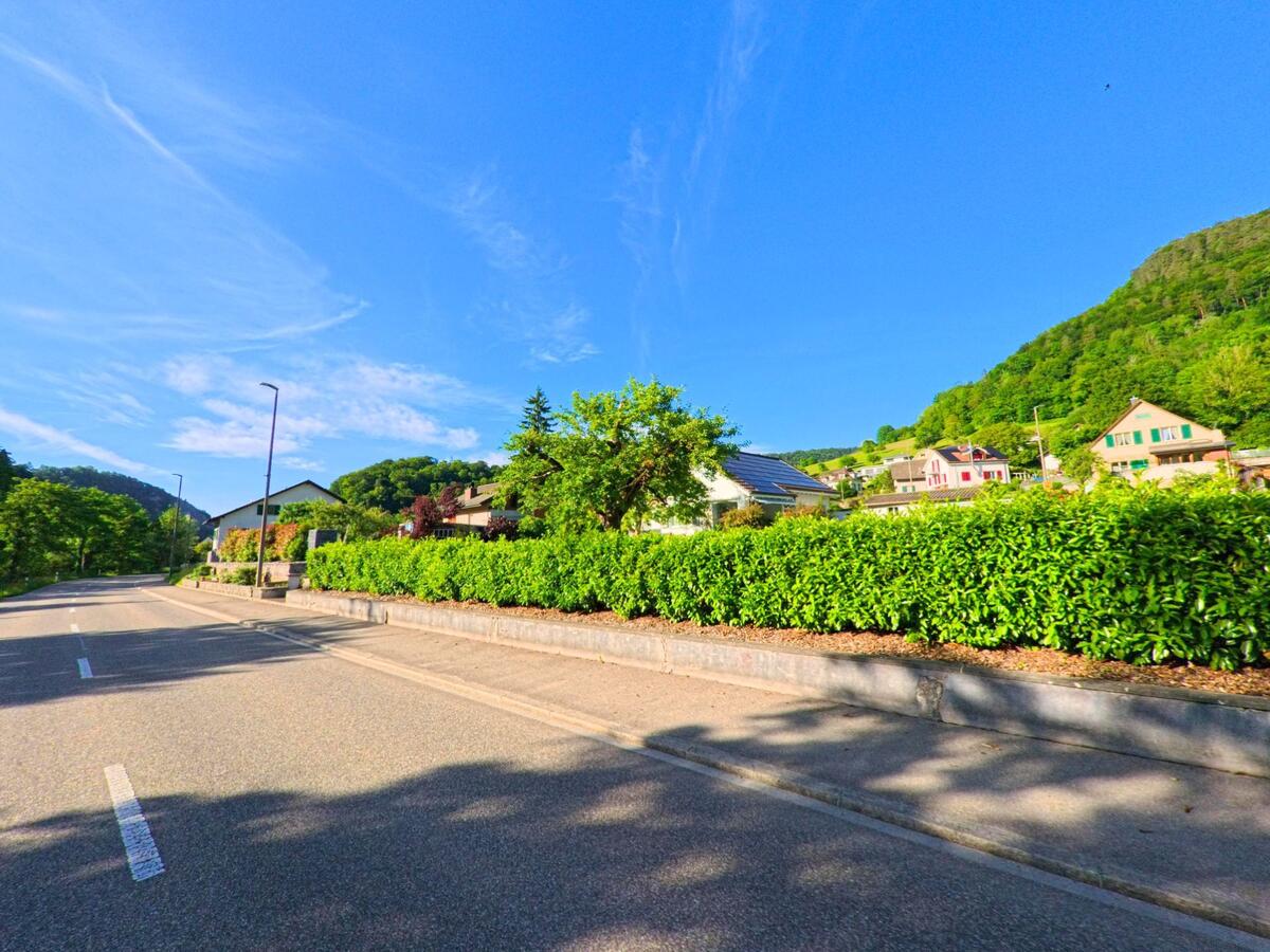 Eine breite Strasse mit Randbepflanzung und modernen Wohnhäusern zieht sich durch das sonnige Erschwil (4228, Kanton Solothurn) am Fuss der bewaldeten Hügel entlang.