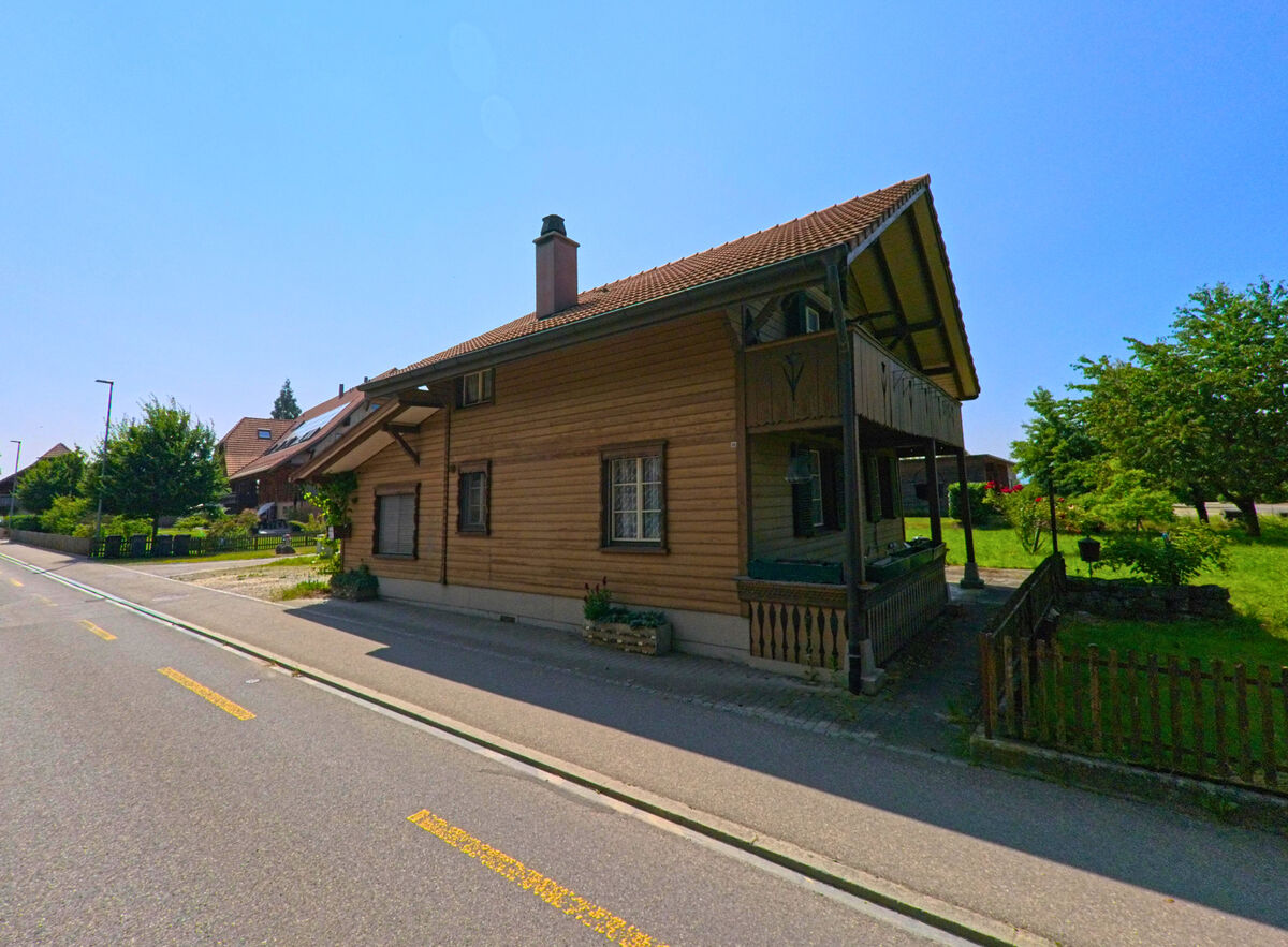 In Ersigen (3423, Kanton Bern) steht ein schmales Holzhaus mit überdachter Veranda direkt an der Strasse, umgeben von einer grünen Wiese.