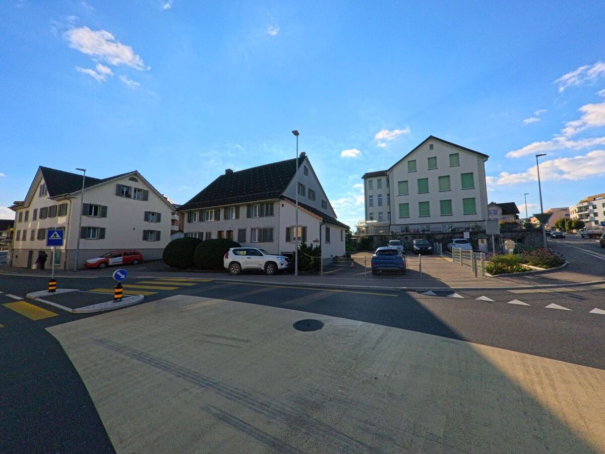 An einer verkehrsberuhigten Kreuzung in Eschenbach (8735, Kanton St. Gallen) stehen mehrere helle Wohnhaeuser mit Parkplaetzen und Zebrastreifen unter blauem Himmel.