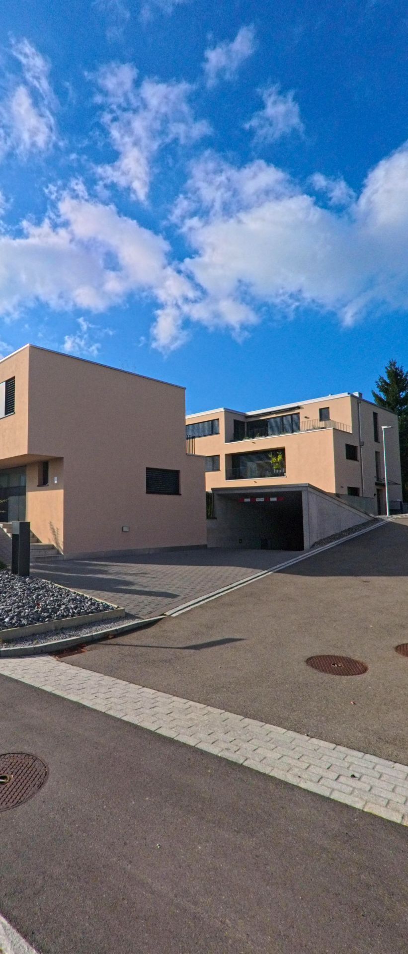 Neben einer Einfahrt zu einer Tiefgarage stehen in Eschenbach (8735, Kanton St. Gallen) moderne Wohngebaeude mit klaren Linien unter einem weitlaeufigen Himmel.