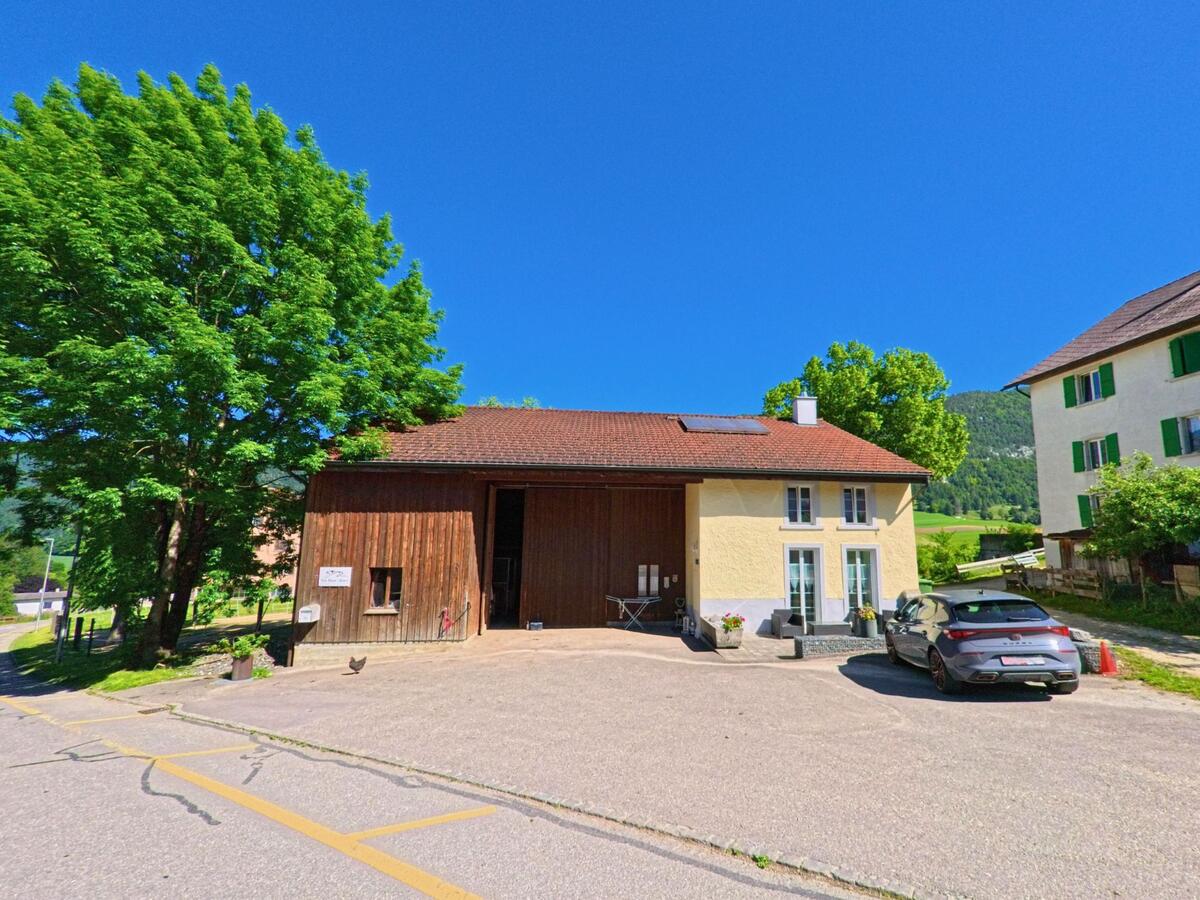Vor einem gelb verputzten Wohnhaus mit angebautem Holzschopf in Eschert (2743, Kanton Bern) stehen ein Auto und Blumenkübel, während im Hintergrund grüne Felder sichtbar sind.