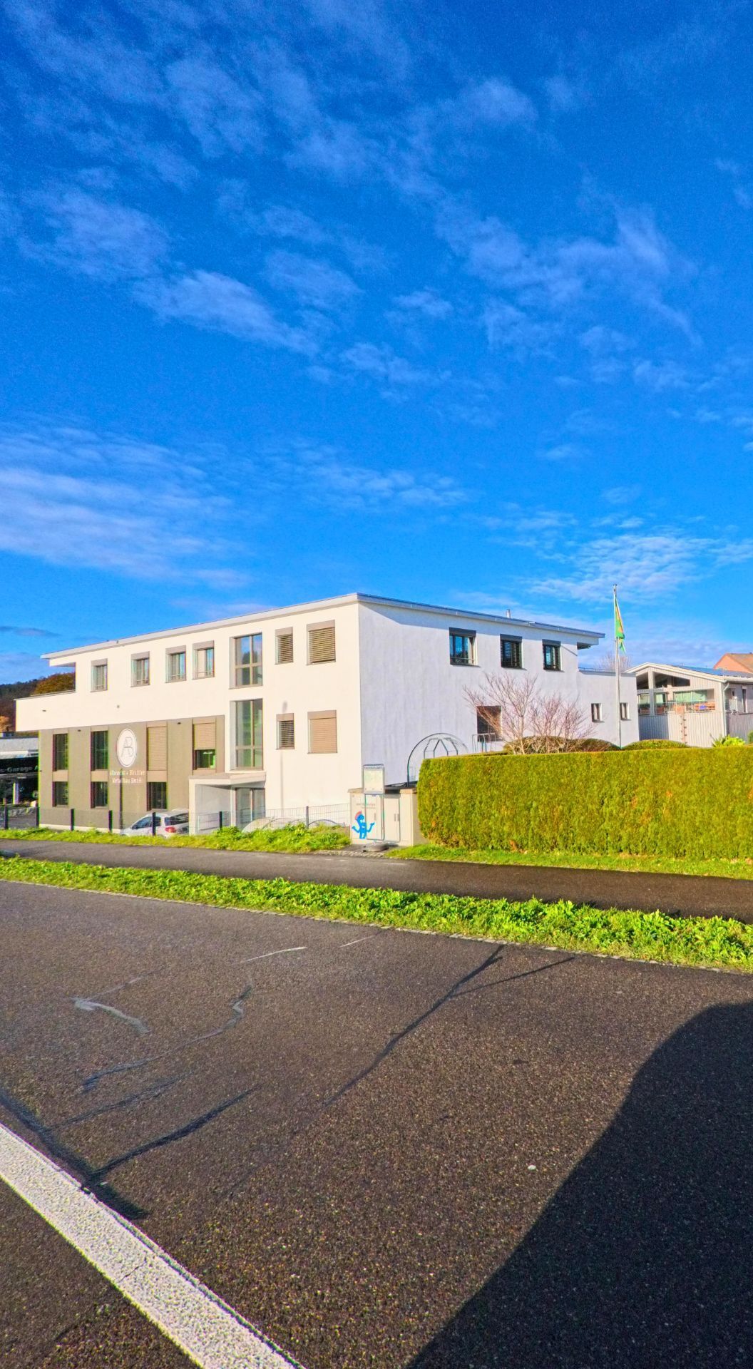 Vor einer Hecke und einem Gehweg steht in Eschlikon (8360, Kanton Thurgau) ein modernes, helles Gebäude unter weitem Himmel mit leichten Wolken.