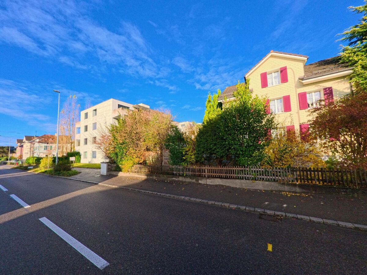 An einer Quartierstrasse in Eschlikon (8360, Kanton Thurgau) stehen ein Haus mit roten Fensterläden und ein modernes Mehrfamilienhaus hinter dichtem Grün und einem Gartenzaun.