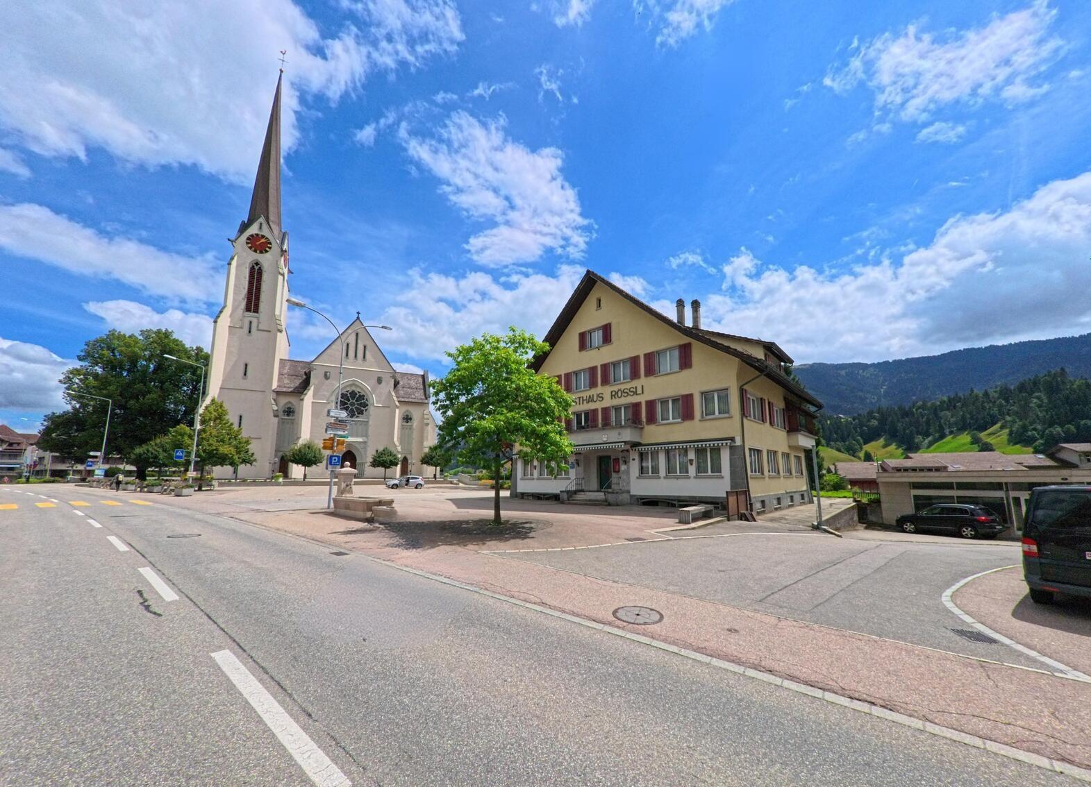 Am Dorfplatz in Escholzmatt-Marbach (6182, Kanton Luzern) stehen eine Kirche mit hohem Turm und ein traditionelles Gebäude an einer Strassenkreuzung vor grünen Hügeln.