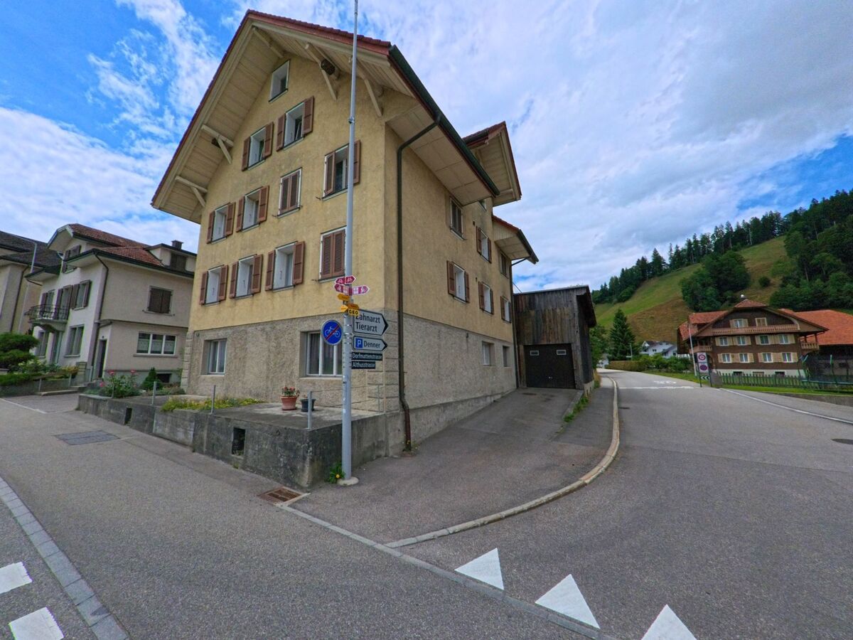 Ein auffälliges Mehrfamilienhaus mit Fensterläden steht an einer Weggabelung in Escholzmatt-Marbach (6182, Kanton Luzern), umgeben von Wegweisern und sanften Hügeln.