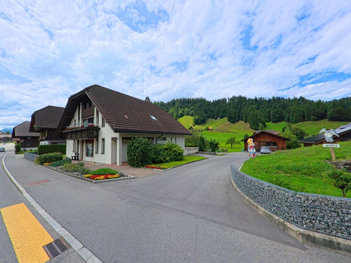 In Escholzmatt-Marbach (6196, Kanton Luzern) steht ein gepflegtes Einfamilienhaus mit Balkonblumen am Rand eines ruhigen Wohnquartiers, dahinter erstrecken sich grüne Hügel und Wald.