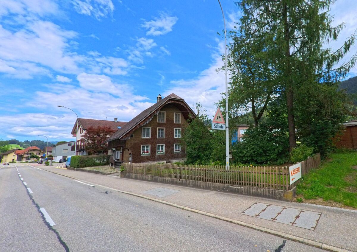 An einer Durchgangsstrasse in Escholzmatt-Marbach (6196, Kanton Luzern) steht ein traditionelles Holzhaus neben einem Schild, das auf eine nahegelegene Schule hinweist.