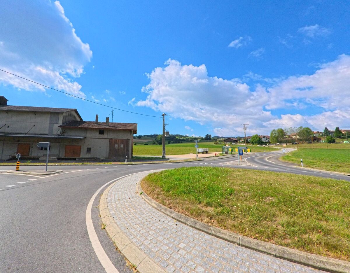 Am Ortsrand von Essertines-sur-Rolle (1186, Kanton Waadt) führt eine Strasse mit Bushaltestelle und Feldern an einem grossen Industriegebäude vorbei.