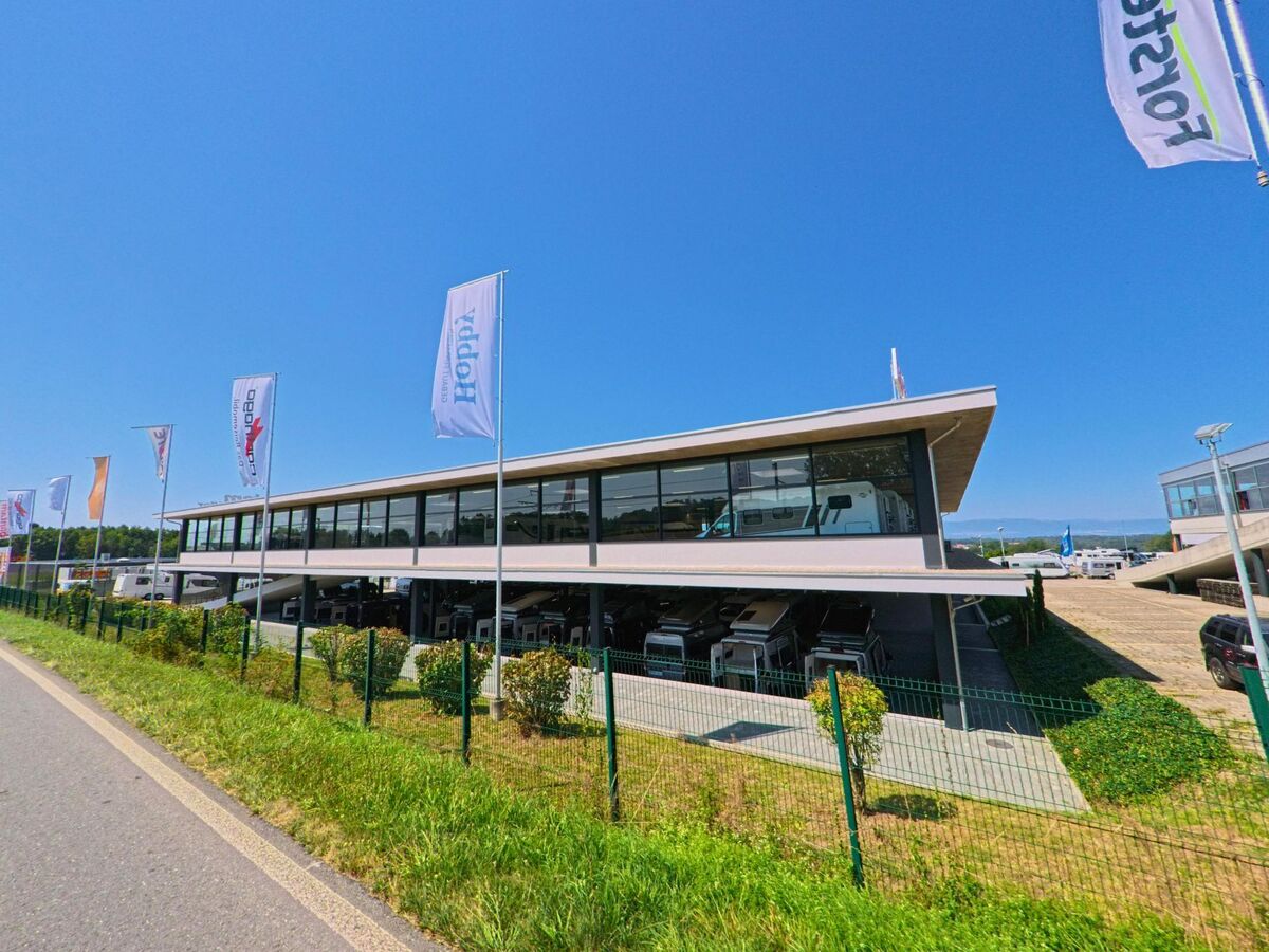 Ein modernes Ausstellungsgebäude mit grossen Glasfronten in Etagnières (1037, Kanton Waadt) zeigt unter dem Vordach abgestellte Fahrzeuge und wird von Fahnen gesäumt.