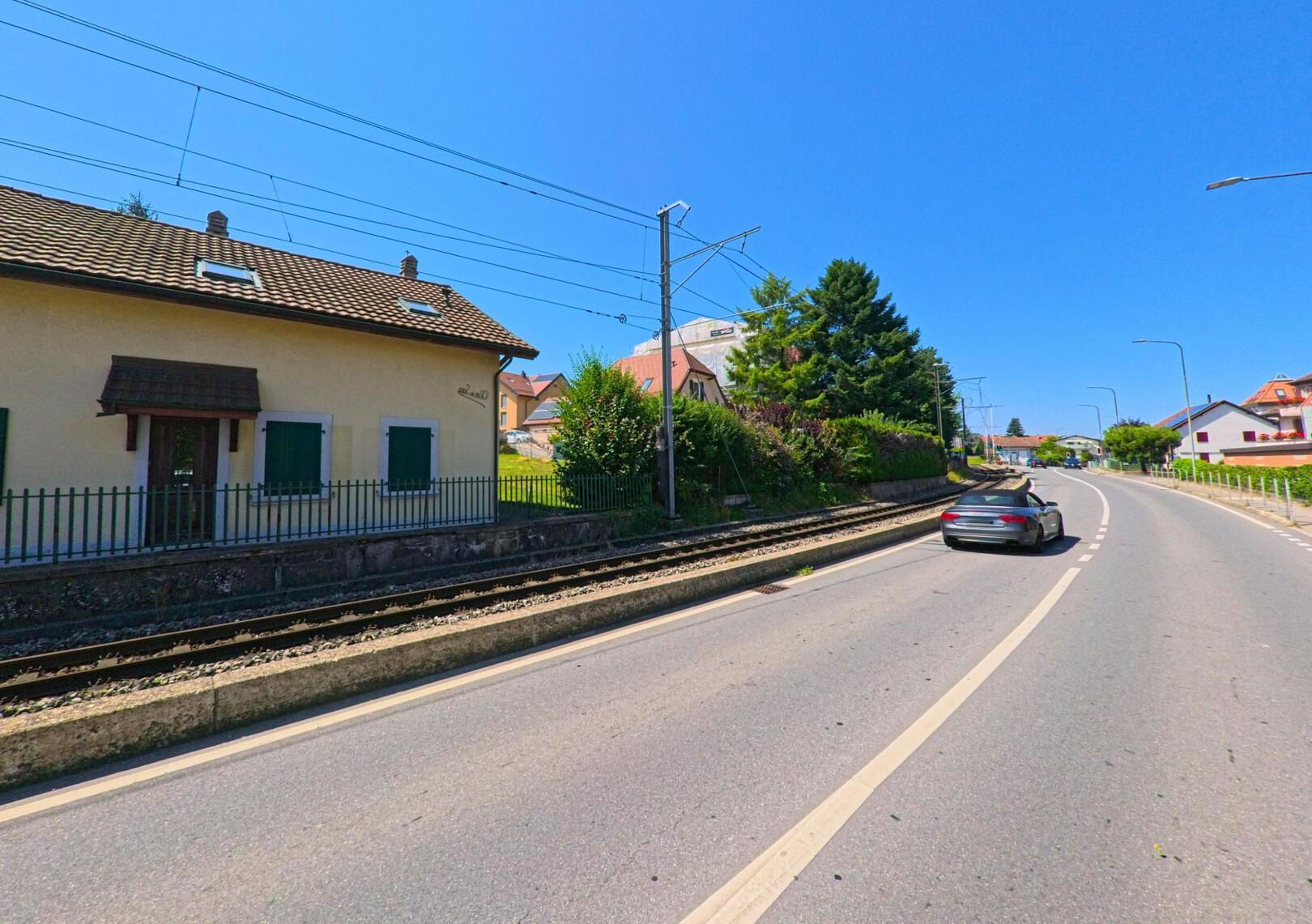 Entlang der Strasse in Etagnières (1037, Kanton Waadt) verläuft eine Bahnlinie mit einem kleinen gelben Haus am Gleisrand.