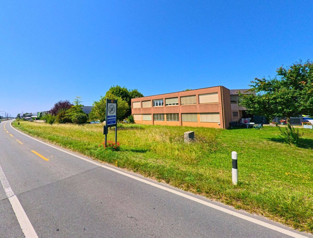 Am Ortseingang von Lausanne nahe Etagnières (1037, Kanton Waadt) verläuft eine Landstrasse mit einem Schild zur Geschwindigkeitsbegrenzung neben einem flachen Gebäude.