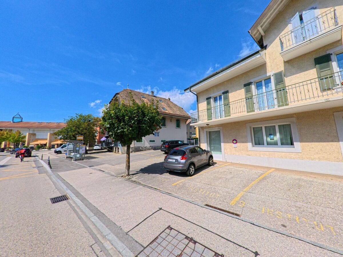 Im Zentrum von Etoy (1163, Kanton Waadt) stehen Wohnhäuser mit Balkonen und ein Platz mit Parkplätzen, im Hintergrund sind weitere Gebäude sichtbar.